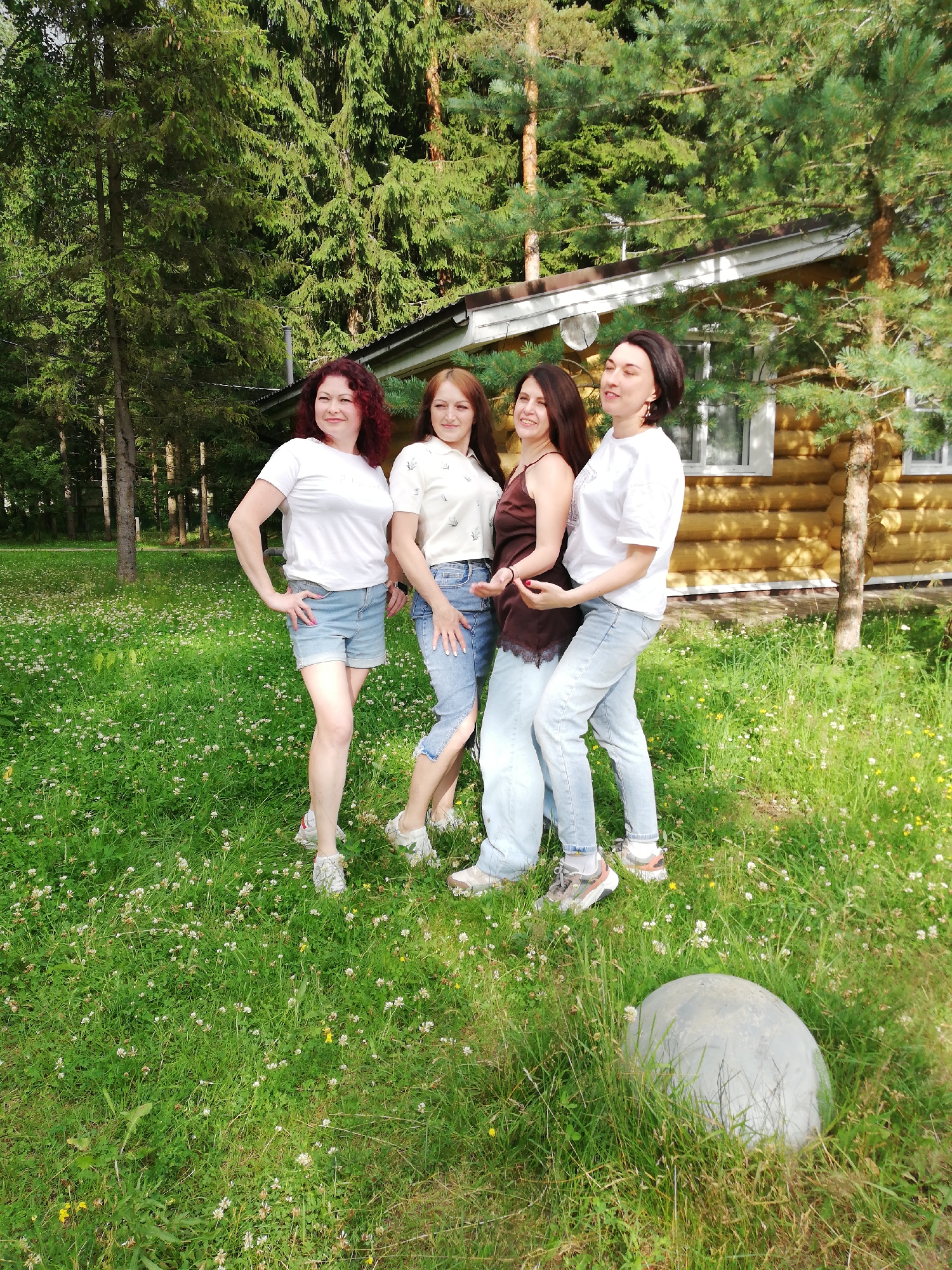 Four women are standing in a grassy area with a log cabin in the background. The woman on the far left has curly red hair and is wearing a white t-shirt and denim shorts. The second woman from the left has straight brown hair and is wearing a light-colored shirt and jeans. The third woman from the left has dark brown hair and is wearing a dark brown camisole and light-colored pants. The woman on the far right has dark brown hair and is wearing a white t-shirt and light-colored pants. The log cabin has a white roof and windows. There are trees in the background. The ground is covered in green grass and white flowers. A large, round, gray object is in the foreground on the right. The image appears to have been taken in the city of Shapki, Russia.
