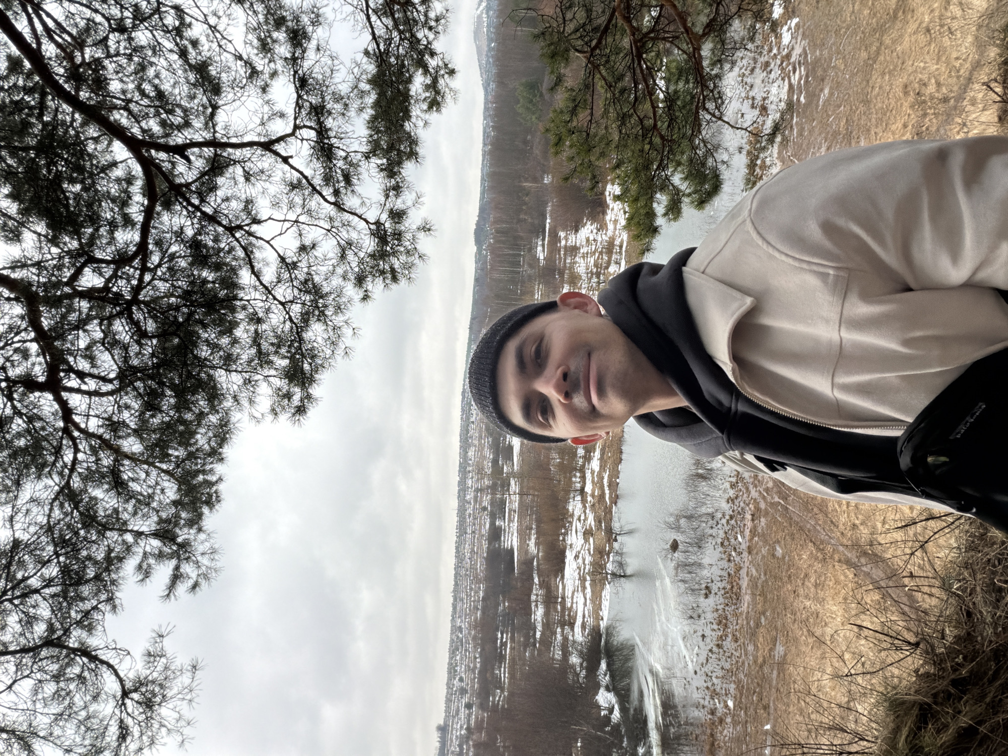 A medium shot shows a person in a beanie, dark-colored hoodie, and beige jacket, standing on a hill. The background reveals a landscape featuring a frozen or partly frozen body of water, possibly a river or lake, adjacent to land covered with a mix of snow and dry vegetation. Beyond this lies a vista of the cityscape, with buildings and structures visible against a cloudy sky. Branches from trees frame the scene on both sides, adding a natural border to the urban and rural elements. The location is likely Izhevsk, Russia.