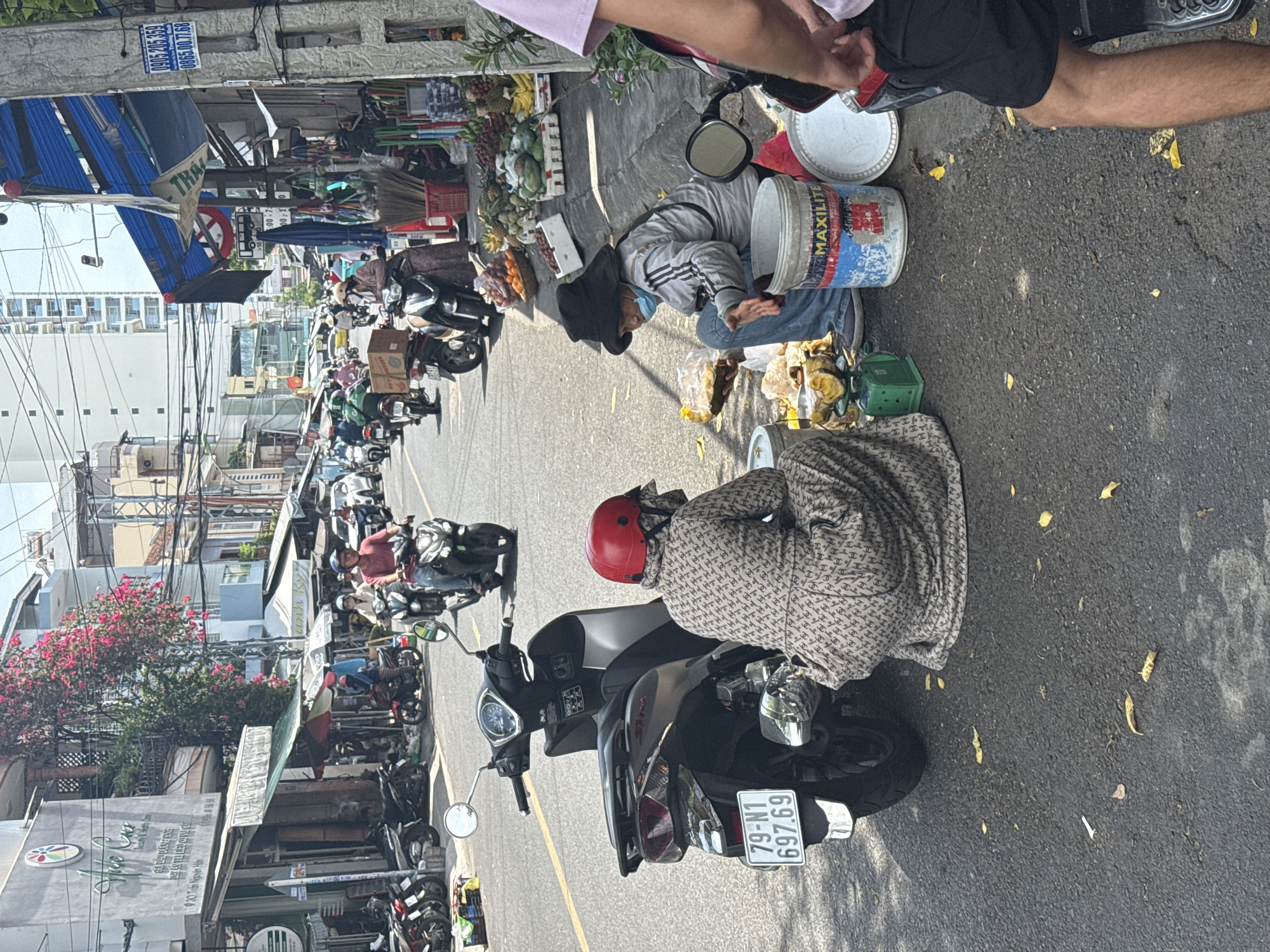 The image depicts a street scene in Nha Trang Ward, Vietnam, likely during daylight hours. The scene is a busy street with a mix of people, vehicles, and market stalls.
In the foreground, two individuals are seated on the asphalt next to motorcycles. One person, wearing a patterned dress and a red helmet, is facing away from the camera. Next to them, another person is wearing a black wide-brimmed hat, a light-colored jacket, blue jeans, and a light blue face mask. This individual is holding a bag of yellow produce and appears to be interacting with the items on a green scale. Several large buckets, one with the label "MAXILITE" visible, and a white lid are also present near the seated individuals. Scattered yellow leaves are on the ground around them.
The street itself is paved and shows signs of wear. On the right side of the image, a pole with advertising stickers and posters is visible, along with hanging goods and market stalls displaying an assortment of fruits and vegetables, including pineapples, papayas, and bananas.
Further down the street, numerous motorcycles are present, both parked and in motion, indicating a common mode of transportation. People are seen riding the motorcycles, some wearing helmets and masks. Buildings line both sides of the street, varying in height and architectural style, with awnings and signs suggesting commercial establishments. In the background, taller buildings are visible, including a multi-story modern structure. Overhead, a complex network of electrical wires is a prominent feature. The overall atmosphere suggests a vibrant urban environment with ongoing daily activities.