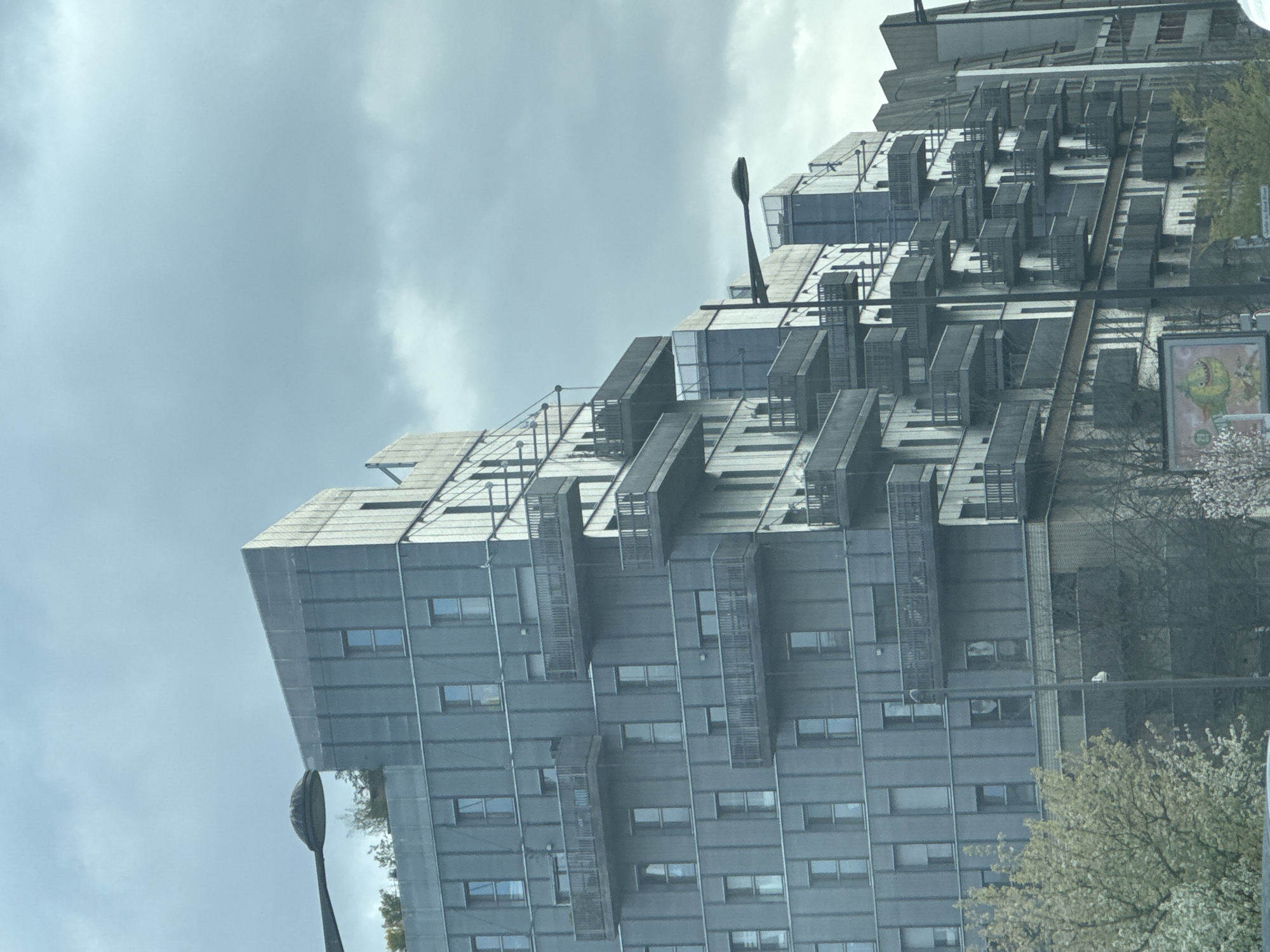 The image shows the Zénith tower in Strasbourg, France. The building features a modern architectural design with a metallic facade, numerous windows, and distinct protruding balconies or extensions. A street light stands in front of the building. A portion of the sky is visible, with overcast conditions. The photograph appears to have been taken from a vehicle. Bare trees and budding blossoms are also visible.