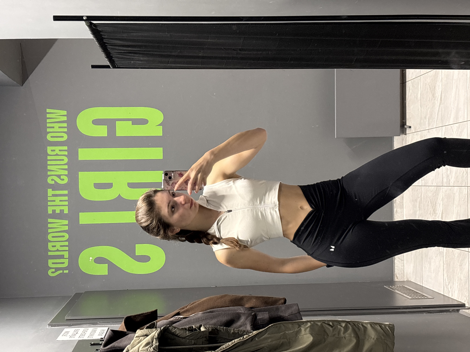 The image shows a woman taking a selfie in a changing room. She is wearing a white zip-up crop top and black leggings. The background features a gray wall with the words "GIRLS WHO RUN THE WORLD?" written in green. There is a black curtain at the top of the frame, likely part of the changing room setup. In the foreground, there are clothes piled on a shelf or bench. The changing room has a somewhat utilitarian aesthetic. It is not possible to determine if this image was taken in the city of Varna, Bulgaria.