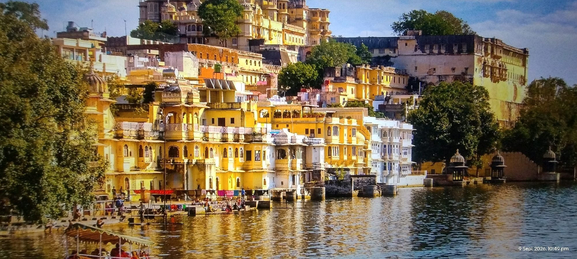 The image depicts a waterfront scene with traditional Indian architecture. In the foreground, a boat with an awning is navigating a body of water, carrying passengers. The water reflects the sunlight and the buildings. Along the shore, there are several multi-story buildings with characteristic Indian architectural elements such as arched windows, balconies, and ornate facades, predominantly painted in yellow and white. People are visible on the waterfront, some engaged in activities like drying clothes, while others are standing or sitting. Lush green trees are interspersed among the buildings and on the higher ground behind them. The background reveals a larger, more imposing structure, possibly a palace or fort, with multiple levels and windows, set against a hazy sky. The overall impression is of a vibrant historical city situated by a lake. The provided information states the location is Yavatmal, India.