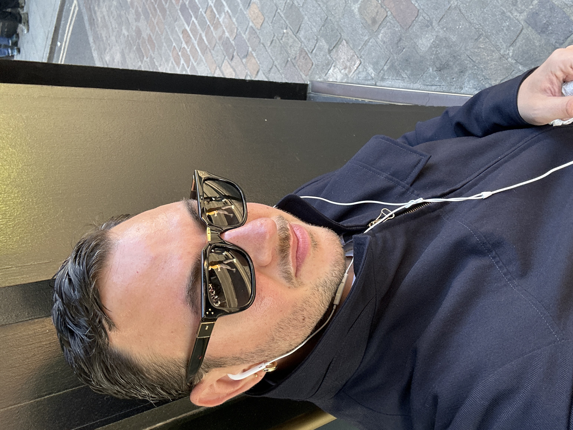 A medium close-up, eye-level shot taken outdoors in bright daylight captures a man resting or reclining. He is positioned horizontally in the frame, suggesting he is lying back on a dark, flat surface, likely a bench or ledge.
The man wears dark, reflective sunglasses with black frames, obscuring his eyes. His hair is short, dark, and appears somewhat styled or wet, and he has a short beard or stubble. He is dressed in a dark blue jacket, possibly denim or a similar sturdy fabric, with a visible zipper down the front and subtle stitching details. White wired earphones are visible, with one earbud inserted into his right ear. In the upper right corner, his hand is holding a crumpled white object, possibly a tissue or paper.
The background reveals a paved street or sidewalk with a textured, brick-like pattern. A faint, light-colored line, potentially a road marking, is visible on the street surface. The reflections in the man's sunglasses offer a blurred glimpse of urban architecture or surroundings. The overall scene is consistent with an urban setting in London, United Kingdom, given the location context. The bright lighting indicates a clear day. The man appears calm and relaxed. Tiny, gold-colored decorative elements or a logo are subtly visible on the arms of his sunglasses.