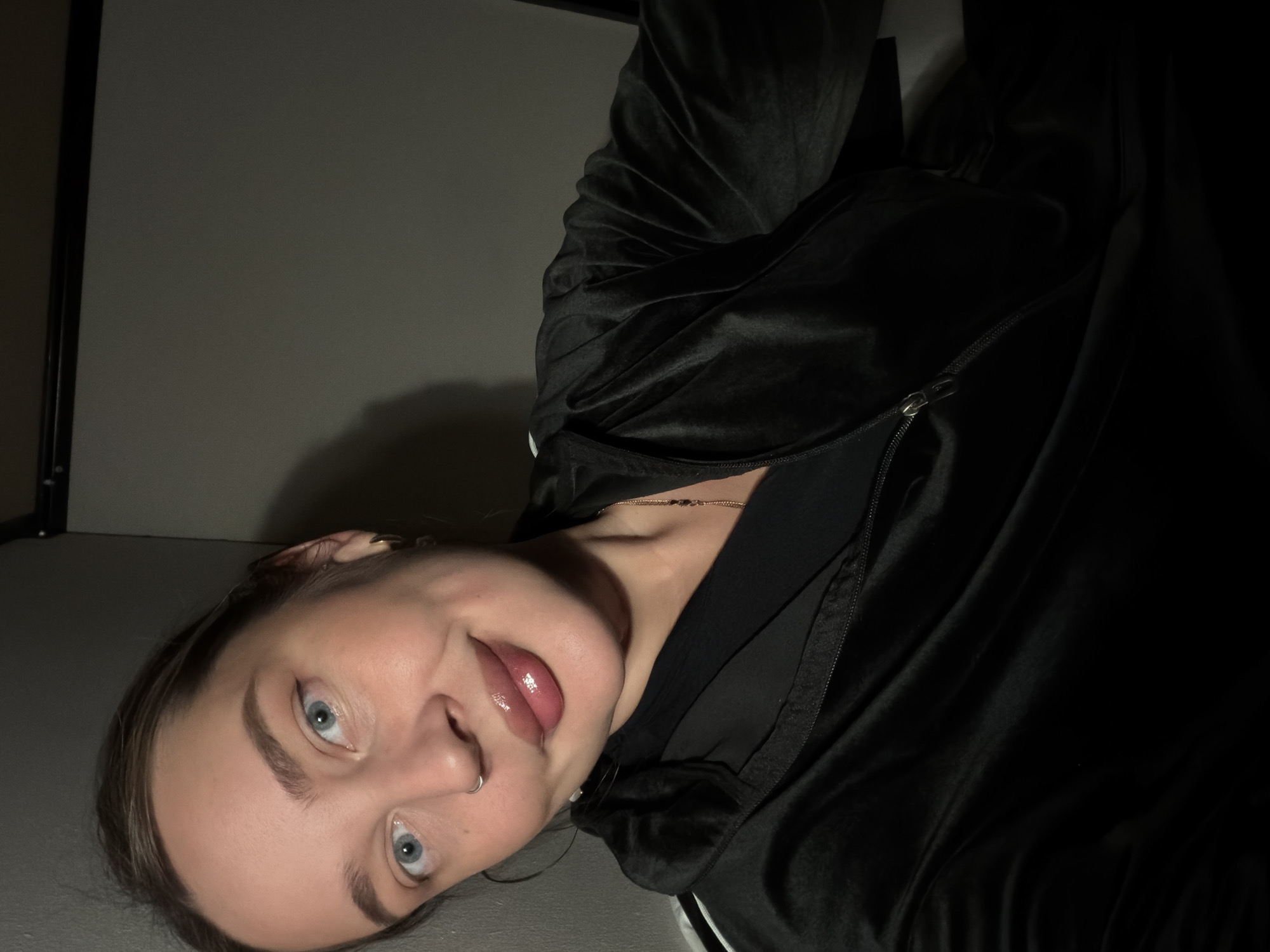 A person is lying on their side on a gray surface. They have light skin, light blue eyes, and their tongue is slightly protruding from their mouth. They are wearing a black, possibly velvet, zip-up jacket and a necklace. A nose ring and hoop earring are visible. The lighting is dim, with a light source appearing to be above and to the left of the person. A section of a black doorframe is visible in the upper-left corner. The location is not Moscow, Russia.