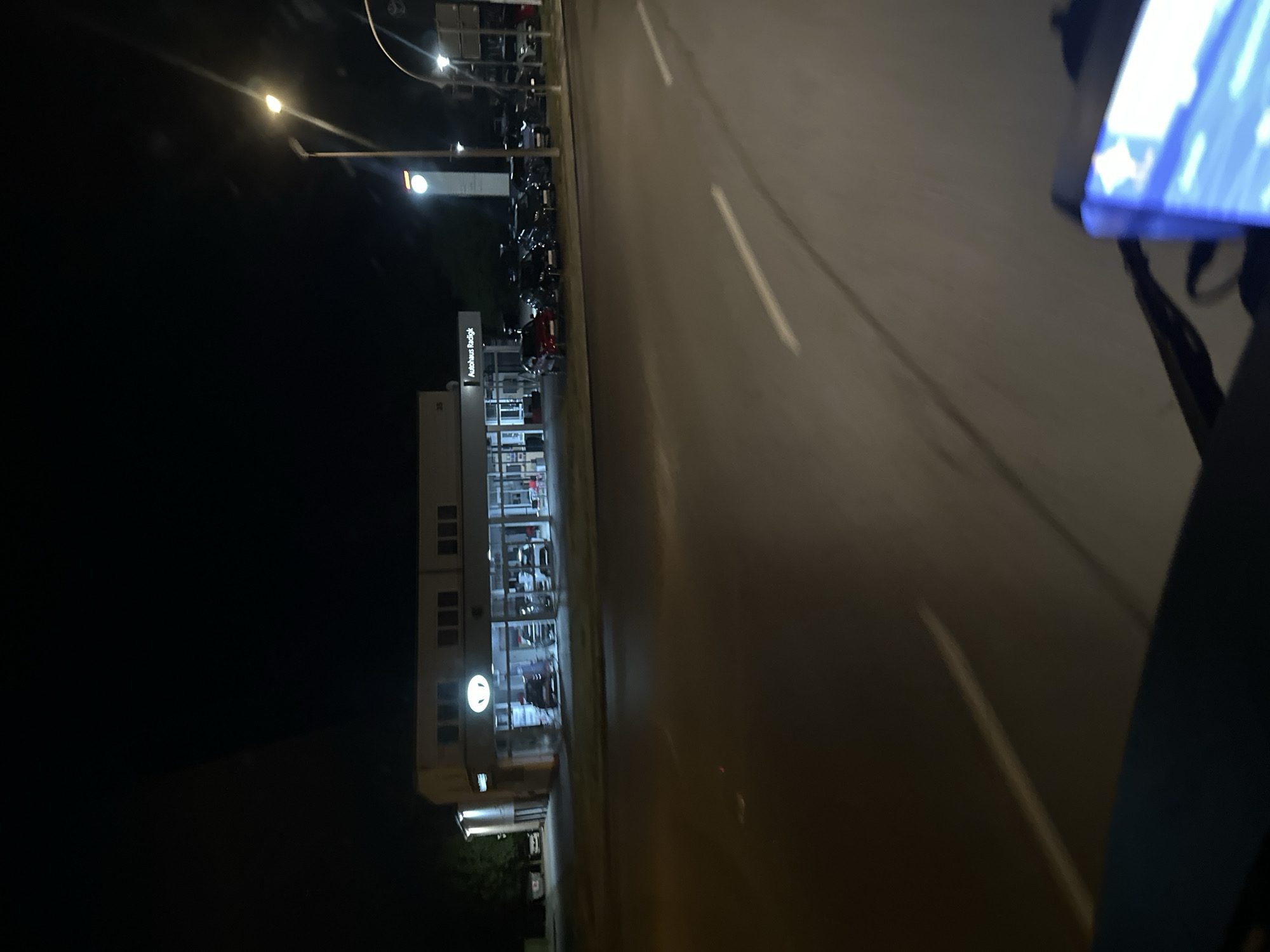 The image depicts a car dealership named "Autohaus Radigk" at night in Lübben (Spreewald), Germany. The dealership building is well-lit, showcasing several cars both inside the showroom and parked outside. A street lamp illuminates the area. The perspective suggests the photo was taken from a moving vehicle, indicated by the motion blur on the road and the edge of a dashboard or screen in the foreground. The overall scene is one of commercial activity continuing into the evening.