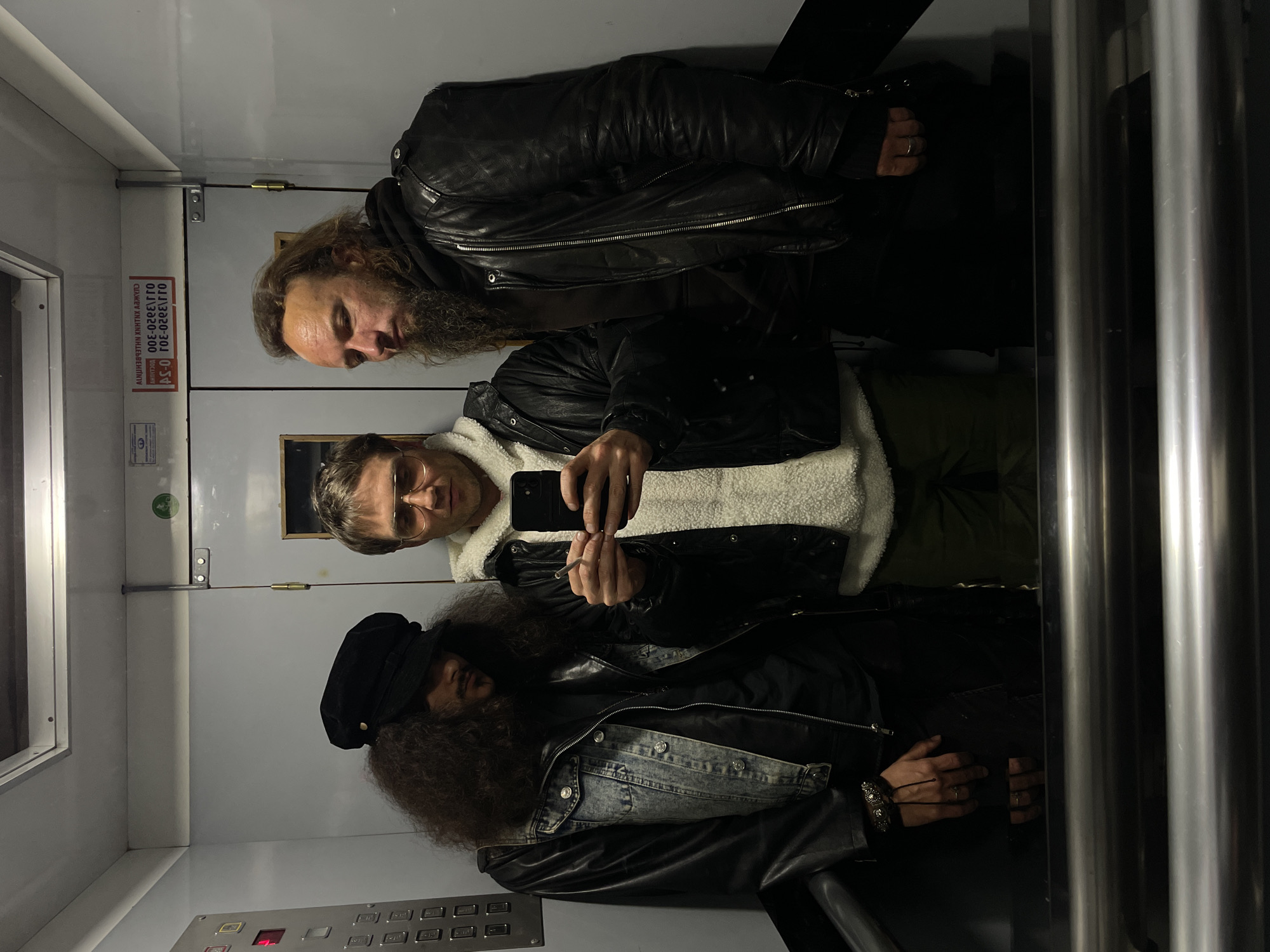 Three men are in an elevator in Belgrade, Serbia. The elevator has gray walls and a metal control panel. The man at the top has long brown hair, a beard, and is wearing a black leather jacket. The man in the middle is wearing round glasses, a white sherpa hoodie, and a black jacket, and is holding a phone and a cigarette. The man at the bottom has long curly hair and is wearing a black cap, a denim jacket, and a leather jacket.
