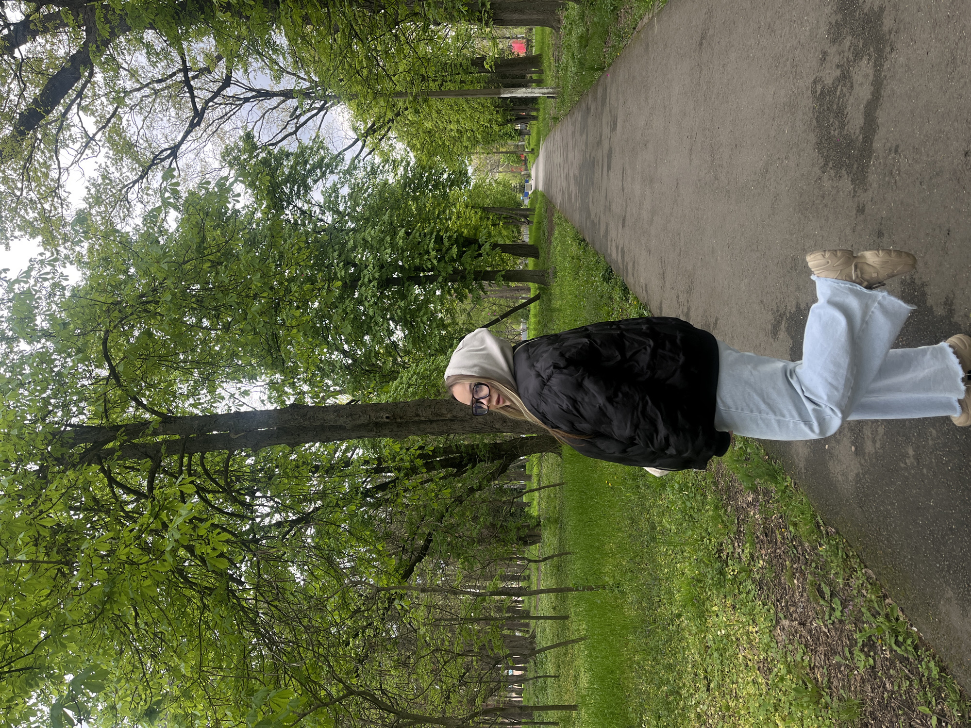 This image depicts a young woman standing on a paved path in a park or tree-lined avenue, likely in Krasnodar, Russia. The woman, positioned on the left side of the path, looks directly at the camera with a neutral expression. She has long, straight blonde hair and wears dark-rimmed glasses. Her outfit consists of a light-colored hooded sweatshirt beneath a black puffer jacket, light blue wide-leg jeans, and chunky beige sneakers.
The setting is characterized by abundant natural elements. To the woman's left, the ground is covered with grass and some patches of bare earth or low-lying vegetation. To her right, a paved path, appearing to be asphalt or concrete, extends into the distance, bordered by more grass. Numerous trees with lush, bright green leaves line both sides of the path, suggesting it is spring or early summer. The tree canopy overhead is dense, with a bright, overcast sky visible through the foliage. In the background, the path recedes into a dense green vista of trees, and a faint red object, possibly a distant building, can be glimpsed through the leaves.
The lighting indicates daytime, with soft, diffused light suggesting an overcast sky or conditions without direct sunlight. There is no visible text in the image.