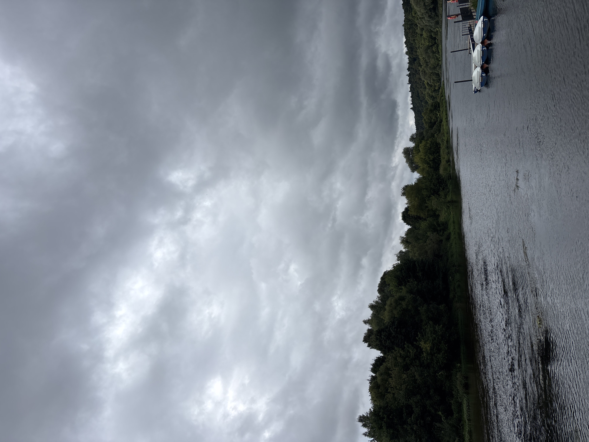 The image shows a river in Zvenigorod, Russia, under a cloudy sky. The river is dark and has ripples, suggesting a gentle current. On the right bank, there is a pier with several small boats docked. The boats are white with blue trim and appear to be recreational. Behind the pier, there is a wooden structure and a hint of orange life vests. The left bank of the river is lined with dense green trees, forming a natural landscape. The sky is overcast with heavy gray clouds, creating a somber mood. The overall scene depicts a natural setting by the river, possibly a recreational area, with an impending sense of rain.