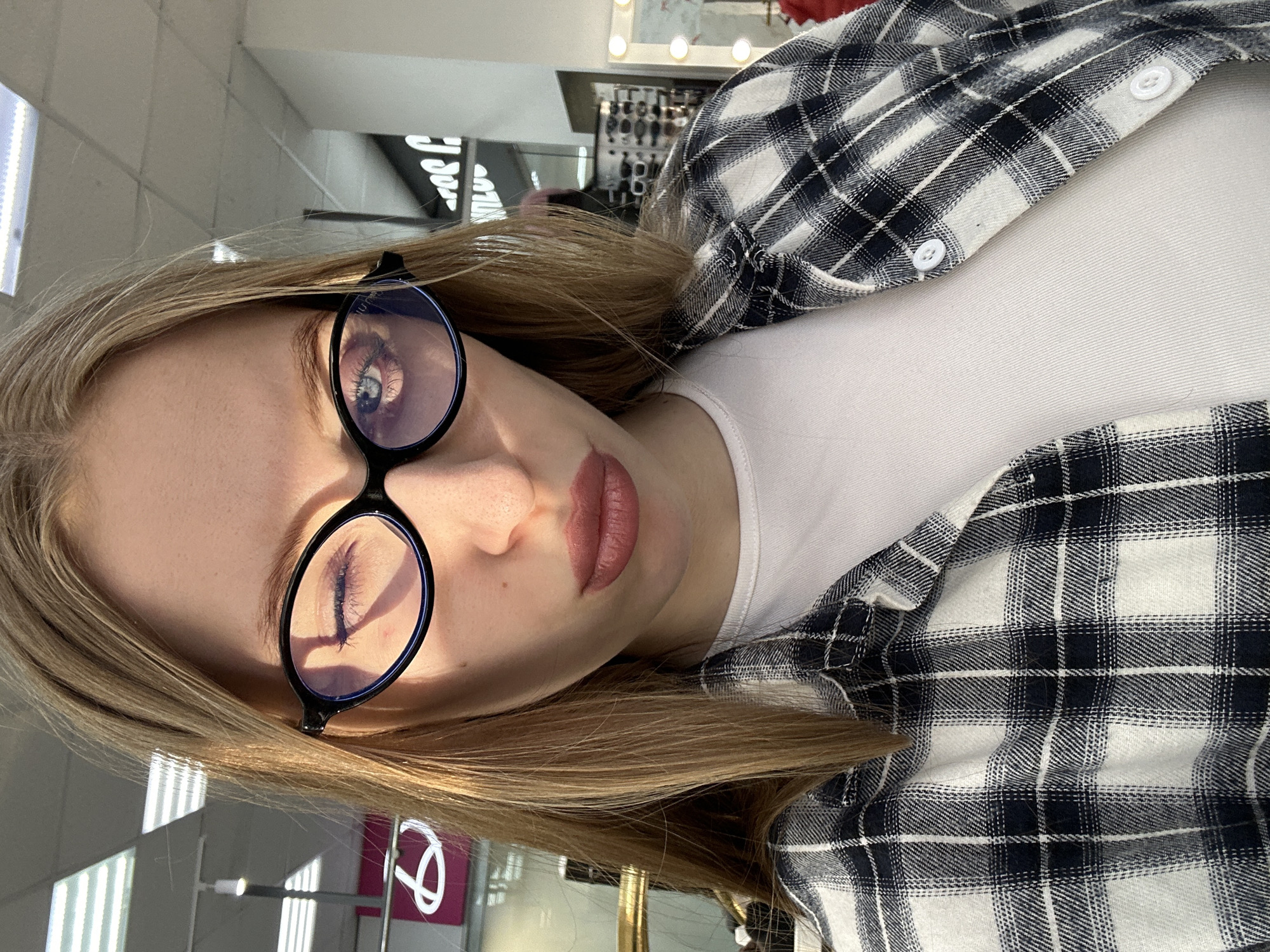 A young woman is depicted in a head-and-shoulders shot within an indoor commercial establishment located in Chaikovsky, Russia. She has light brown hair. Her face is centered, and she is wearing black, oval-framed glasses with a blue tint to the lenses. Her right eye (viewer's left) is closed in a wink, while her left eye (viewer's right) is open and blue, displaying a reflection of an overhead fluorescent light fixture. Her lips are pursed and have a matte pink application. She is dressed in a white crew-neck t-shirt underneath an unbuttoned black and white plaid flannel shirt. The background includes bright, rectangular fluorescent ceiling lights, a blurry illuminated pink logo on the left, and on the right, racks displaying eyewear and a mirror with spherical lights around its perimeter, consistent with an optician's or retail store. Shadows from her glasses frame are visible on her upper face.