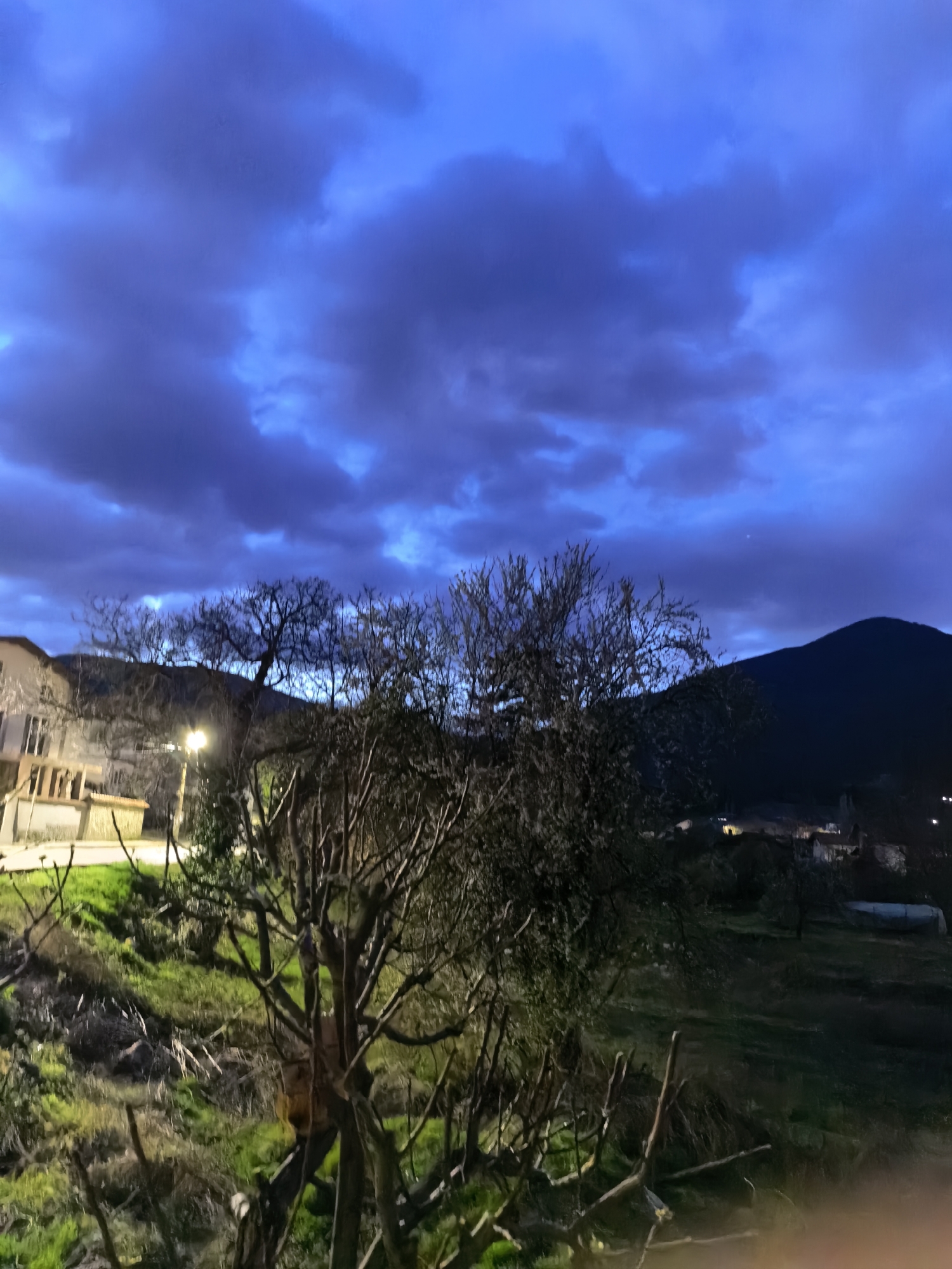 The image depicts an outdoor scene, likely at dusk or dawn. The sky is a deep blue, filled with textured clouds. In the foreground, there is a leafless tree with dark branches. The ground is green, with patches of dried grass. In the mid-ground, a building is visible to the left, illuminated by a street light. A dark, silhouetted mountain is present in the background on the right side of the frame. Faint lights suggest a settlement at the base of the mountain. The location is Unknown, Unknown.