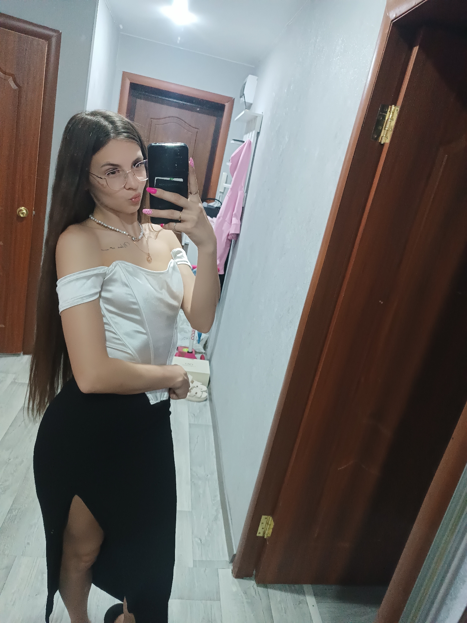 A full-length indoor shot shows a young woman holding a mobile phone up and taking a selfie in a hallway of a building located in Unknown, Unknown. She is positioned facing forward and angled towards the viewer’s left, with a large mirror on the right side of the frame. The woman has long, straight brown hair and wears glasses, a white off-the-shoulder top, and a long black skirt with a thigh-high slit on the left side. She is wearing a pearl necklace and another necklace with a pendant. The phone in her hand has pink dots on the fingernails holding it. Her expression is a slight pout.
Behind her, the hallway is visible, with doors on either side and a glimpse of a room with clothing hanging on a rack. The walls are light-colored. The doors are brown with brass-colored knobs and hinges. The floor is made of light-colored tile. The lighting is bright and appears to be artificial.