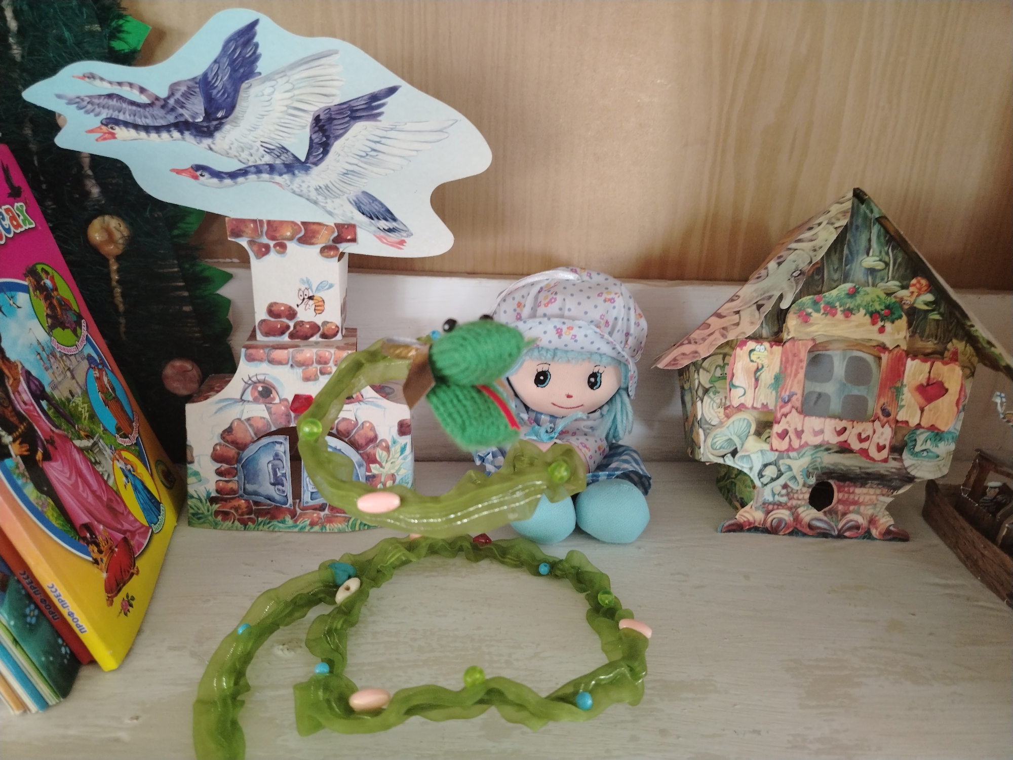 The image depicts a collection of whimsical, handcrafted items displayed on a light-colored wooden shelf. On the left, a stack of colorful books with illustrations of fairy tales and fantastical scenes is leaning against a dark green, artificial Christmas tree. Above the books, a cutout decoration features three geese in flight against a pale blue sky, mounted on a miniature brick structure with a beehive and a buzzing bee.
Dominating the center is a soft doll with blue yarn hair, wearing a floral-patterned bonnet and a blue and white checkered outfit. The doll's head is tilted, and its arms are visible, one of which is entangled with a translucent green, flexible toy shaped like a segmented worm or vine, adorned with small pink and blue beads.
To the right of the doll is a miniature, intricately decorated house that appears to be made of paper or cardboard. This structure has a pointed roof adorned with foliage and berries, and its facade features windows with heart-shaped trim and an arched doorway.
The background is a plain, light-brown wooden wall with visible grain. The overall scene suggests a decorative display, possibly in a child's room or a craft fair, featuring handmade toys and storybook-themed elements. The location in Klyuchi, Russia, is not discernible from the visual elements present in the image.
