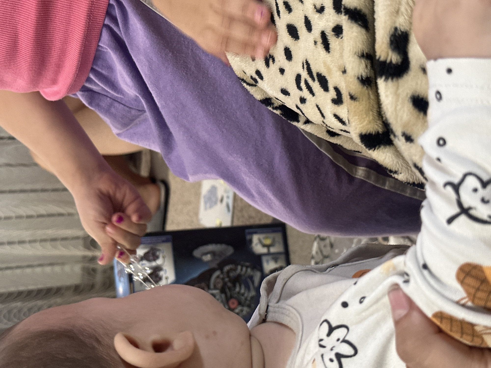 A close-up, slightly angled shot shows a baby lying down. The baby is wearing a white onesie with black and brown patterns. An ear of the baby is visible in the lower left of the frame. Above and to the right of the baby's head is a hand holding a string of small, clear lights. The hand belongs to an individual wearing a pink shirt and purple pants. Another hand is resting on a leopard print blanket, partially obscuring the view. A box with images of what appears to be a game or toy lies near the baby's head. The location is Unknown, Unknown.