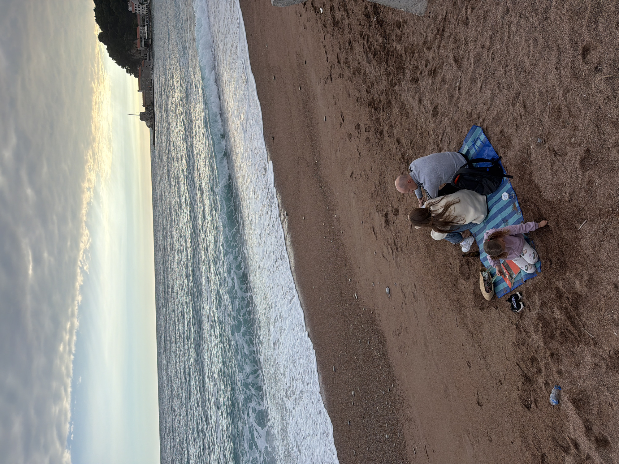 The image shows a beach scene in Petrovac centar, Montenegro. A family of three, including an adult male, a female, and a young child, are gathered on a blue-striped beach blanket. A black backpack and a straw bag are placed on the blanket next to them. The child is reaching for an object. The beach is sandy, and the sea is visible with breaking waves. In the distance, there is a cliff face with buildings on top. The sky is cloudy.