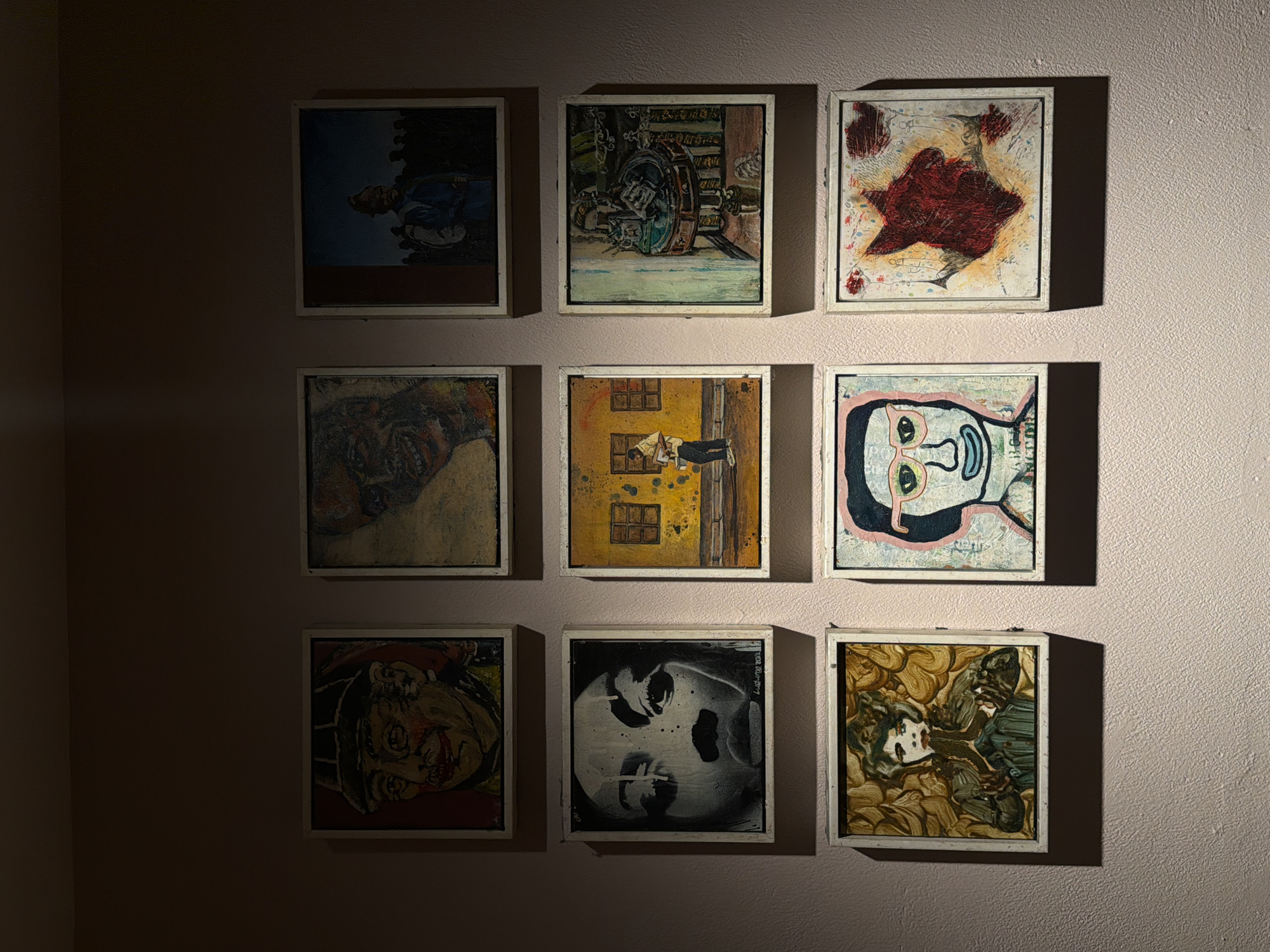A wall is adorned with nine framed paintings, arranged in a 3x3 grid. The paintings vary in style and subject matter, but all feature a white frame. The lighting is uneven, with shadows obscuring parts of the wall and some paintings. The wall is pale pink and textured. The location appears to be an art gallery or a similar display space. The location in London, United Kingdom cannot be ascertained from the provided image.