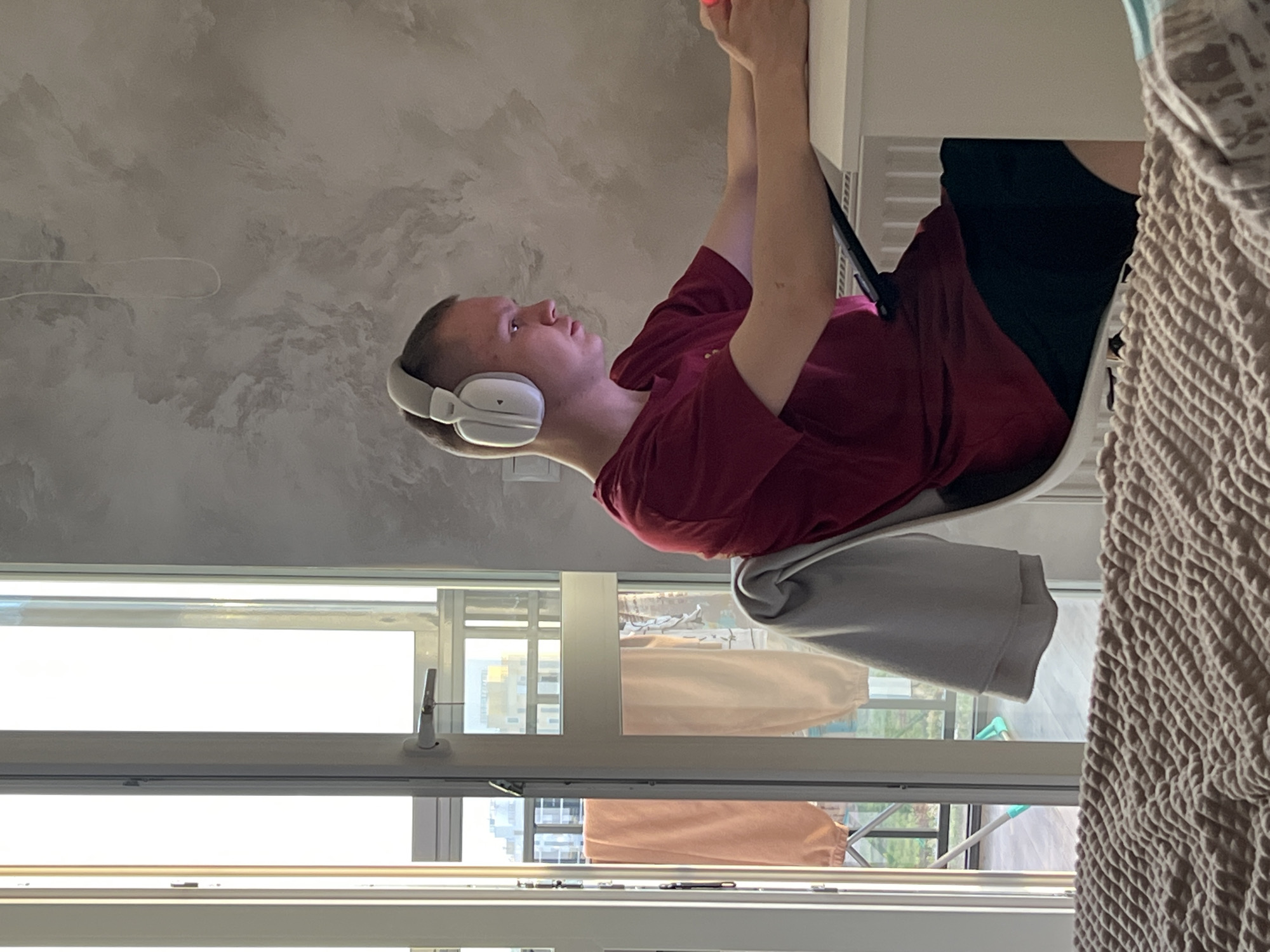 The image shows a person with short blonde hair wearing white headphones and a burgundy t-shirt, sitting in a chair with a grey jacket draped over it. The person is holding their arms up and appears to be interacting with something on a white desk. They are wearing black shorts.
The location is likely an apartment in Izhevsk, Russia, as indicated by the urban scenery visible through the window. The window reveals apartment buildings across a balcony, suggesting an upper-floor location within the city. Drying laundry is visible outside on the balcony. The wall behind the person has a textured, cloud-like pattern. To the right of the person is a beige knitted or woven blanket.