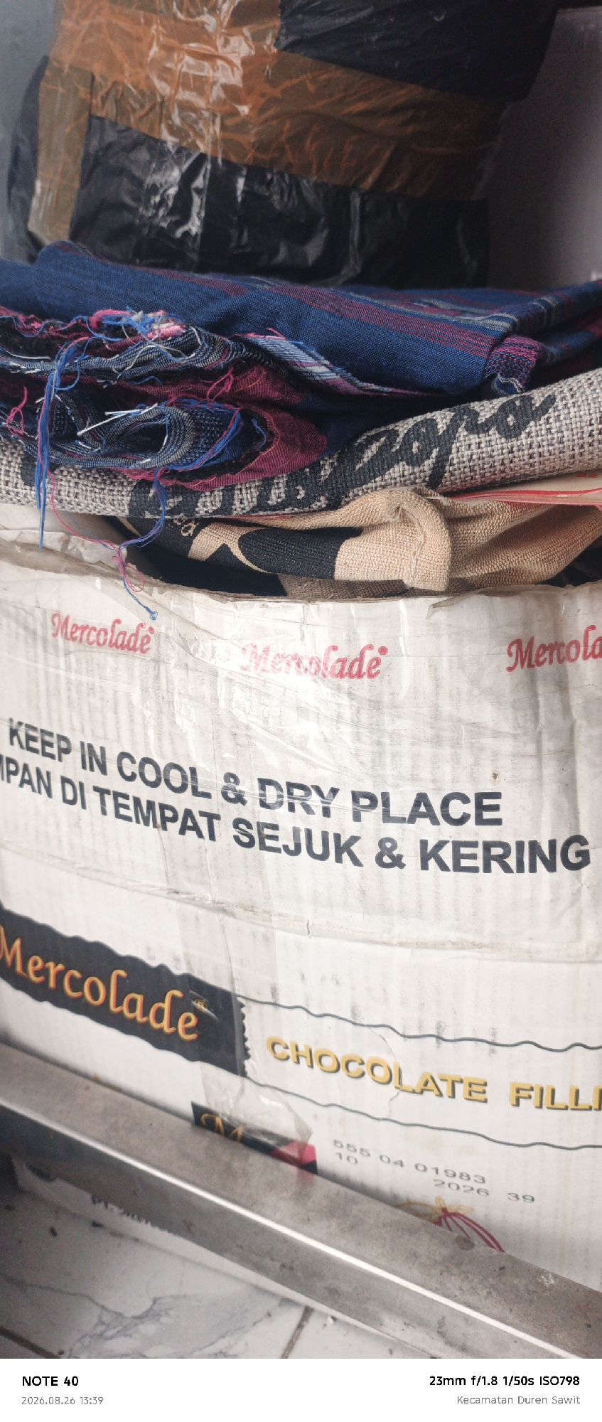 A close-up, eye-level, vertical shot shows stacked items, possibly within a warehouse or storage area in the city of East Jakarta, Indonesia. The foreground features a silver metal railing or shelving unit. Above this, a white plastic-wrapped container or sack is visible, bearing the red text "Mercolade" multiple times and English and Indonesian instructions: "KEEP IN COOL & DRY PLACE" and "MPAN DI TEMPAT SEJUK & KERING". Below these instructions, a horizontal band displays the word "CHOCOLATE FILLI" in yellow text. Further up, several layers of fabric are piled, including a dark blue fabric with red stripes and frayed edges, and a beige fabric with black printed motifs. The uppermost portion of the image shows black plastic packaging secured with orange tape. The bottom of the image displays text overlay identifying the photo as "NOTE 40", taken on "2026.08.26 13:39" with camera settings "23mm f/1.8 1/50s ISO798" and location "Kecamatan Duren Sawit".