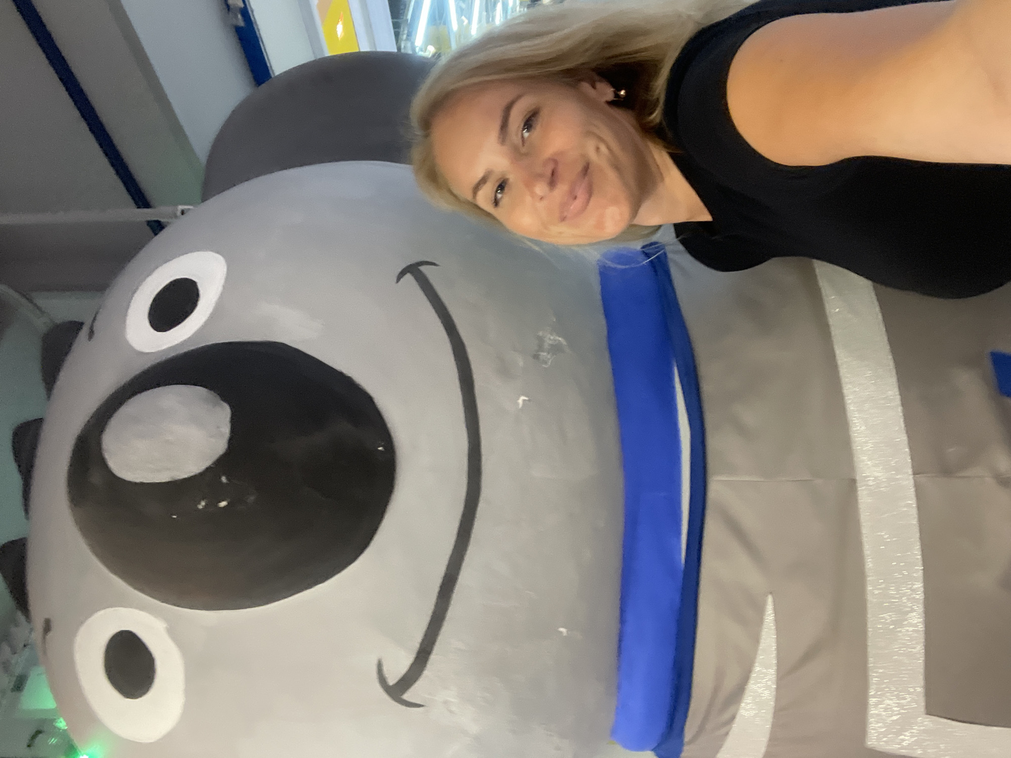 The image depicts a fair-skinned woman with blonde hair and a dark-colored t-shirt posing next to a large, cartoon-like grey character with oversized eyes and a wide smile. The character seems to be wearing a blue collar, and the background suggests an indoor, possibly public, space. The image was taken in Sochi, Russia.