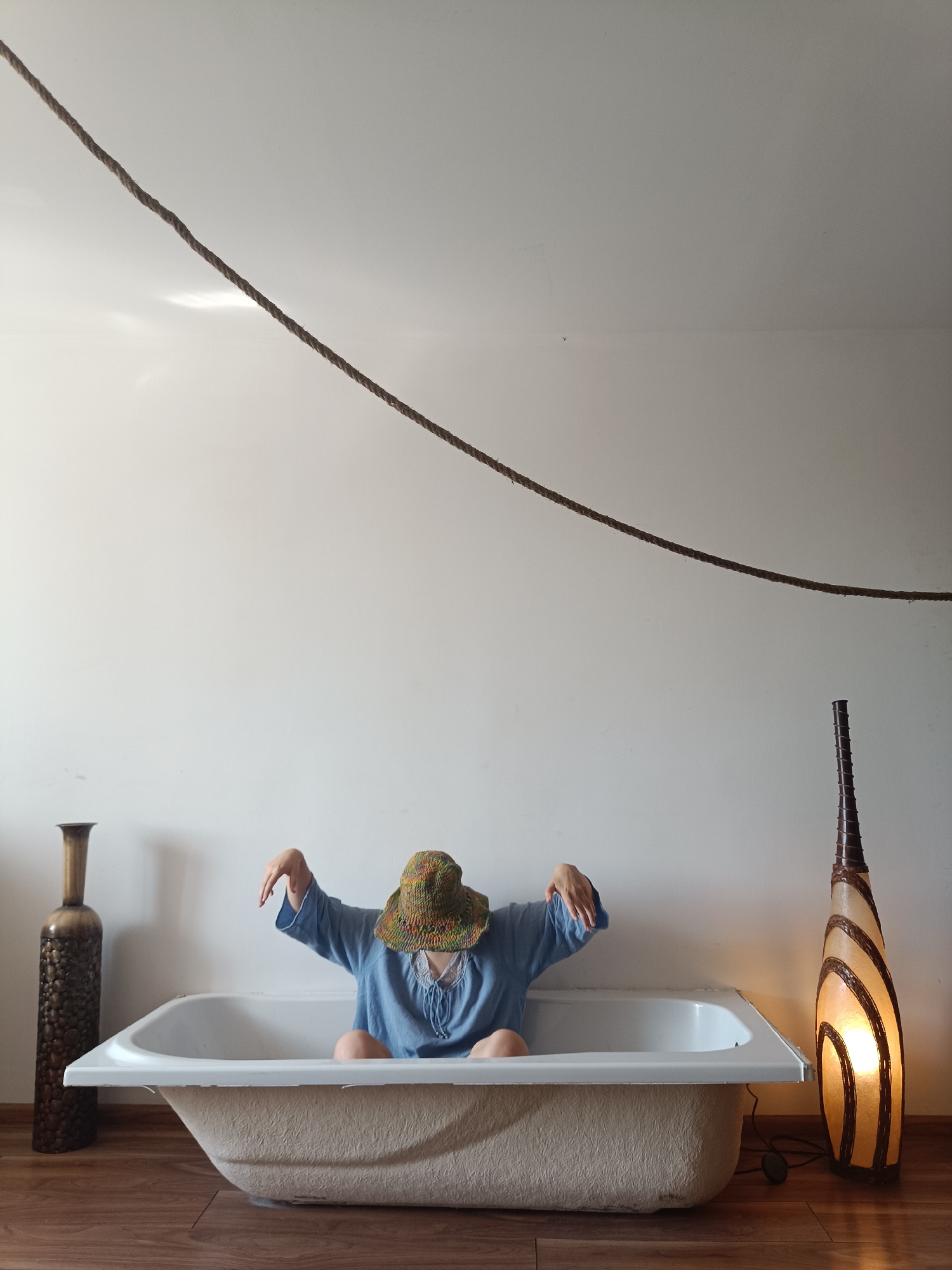 The image depicts an indoor scene, likely a room within a residential dwelling in Łódź, Poland. A person wearing a blue shirt and a colorful woven hat is seated in a white bathtub. Their face is obscured by the hat, and their arms are extended outwards with their hands slightly curled. The bathtub is positioned on a wooden floor against a plain white wall. To the left of the bathtub stands a tall, dark decorative vase with an ornate pattern. To the right, a floor lamp with a distinctive spiral design, illuminated with a warm light, stands near the bathtub. A thick rope is suspended across the upper portion of the frame, diagonally from the top left to the top right. The lighting suggests an indoor environment with diffused natural light and the artificial light from the lamp.
