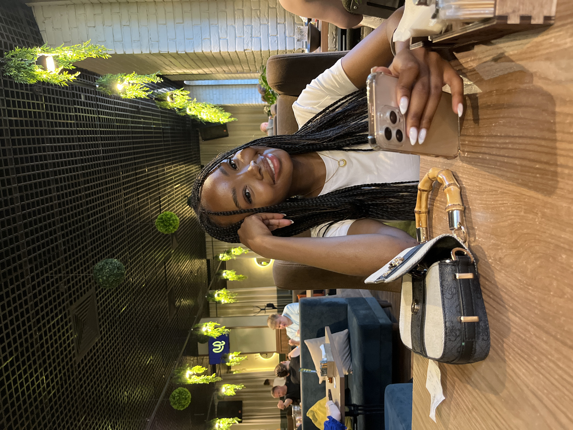 In a Kazan restaurant, a young Black woman with braided hair sits at a wooden table, looking at her phone. She wears a white t-shirt and a gold necklace. A handbag with a bamboo handle rests on the table beside her. The restaurant's interior features a grid ceiling with hanging plants and exposed brick walls. In the background, other patrons are visible.