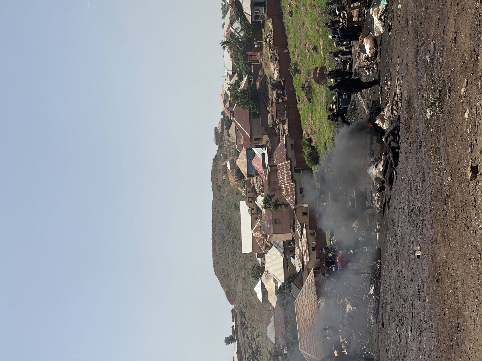 The image shows an open dump site in Jos, Nigeria, with smoke rising from burning waste. The foreground consists of dark, bare earth littered with debris. In the middle ground, a group of people appear to be scavenging or working at the edge of a burning pile. Behind them, a hillside is dotted with residences of varying sizes and construction materials, indicating a dense neighborhood built on an incline. The sky is pale blue, suggesting a clear day.