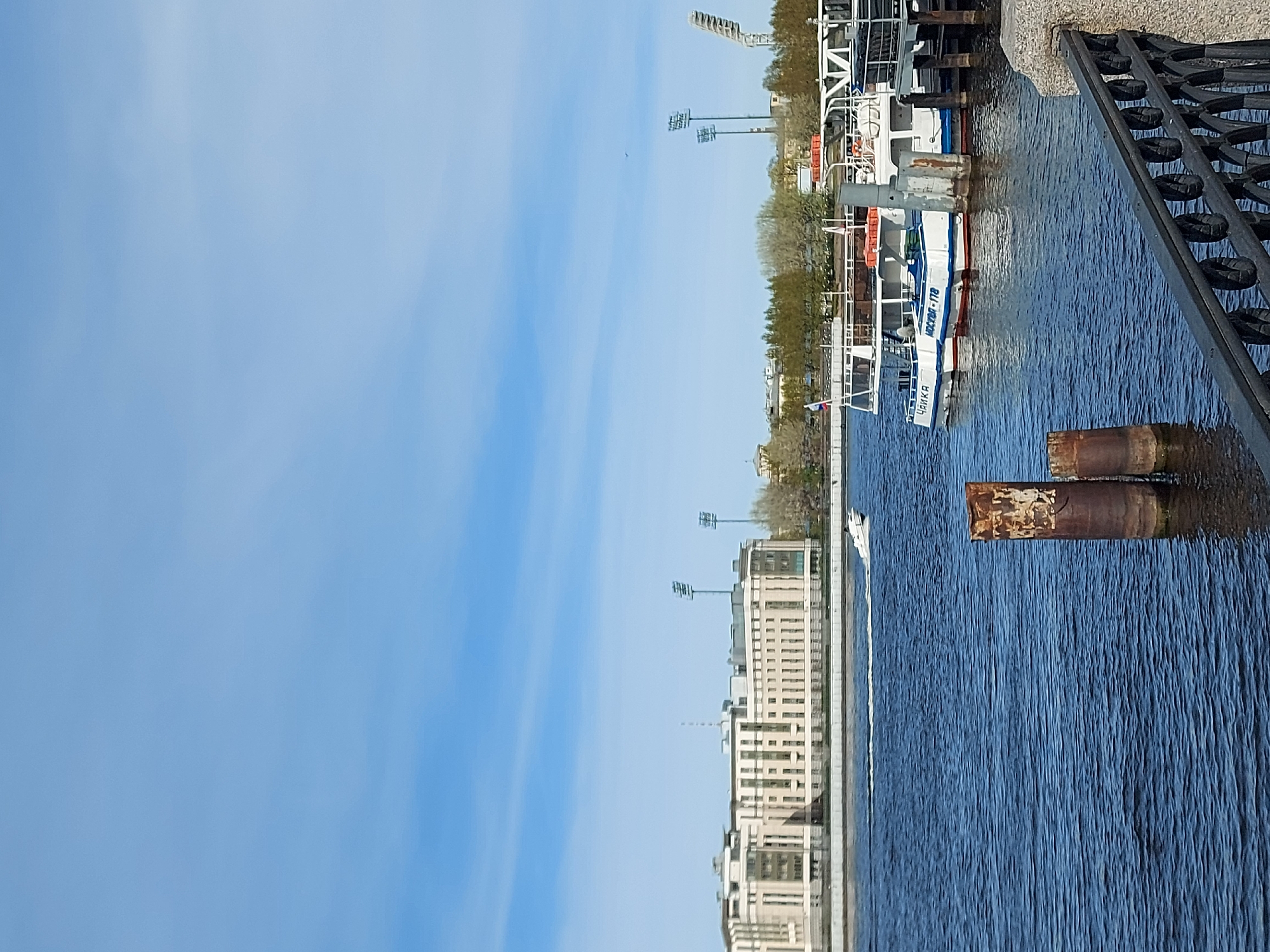 The image depicts a waterfront scene in Saint Petersburg, Russia, featuring the Neva River. A passenger boat, identified as "ЧАЙКА MOCKBA-178," is docked along the embankment, near a metal railing. The river surface reflects the surrounding architecture, including buildings and lamp posts. Two corroded metal posts are partly submerged in the water. The sky is blue, with some wispy clouds.