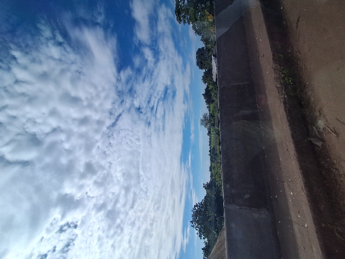 The image depicts a daytime, rural or semi-rural landscape in Kungana, Nigeria. The upper two-thirds of the frame features a blue sky with extensive scattered white cumulus and cirrus-like clouds. A dark gray concrete barrier, appearing weathered, traverses horizontally across the mid-section of the image, showing some surface reflections. The immediate foreground consists of an unpaved dirt surface with sparse green vegetation and scattered debris. Beyond the concrete barrier, a verdant landscape extends into the distance, characterized by numerous green trees, open fields, and faintly visible, partially obscured structures in the background, possibly buildings with light-colored roofs. No individuals or dynamic actions are present in the scene.