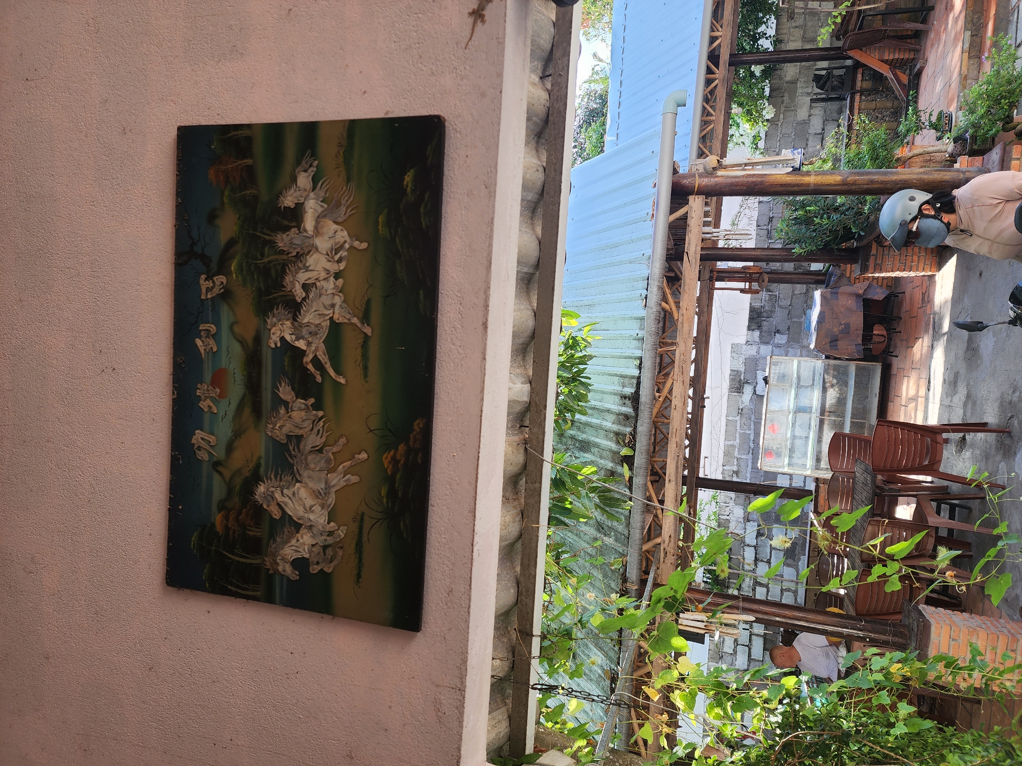 In Phan Thiet Ward, Vietnam, an outdoor dining area is depicted. A painting of eight horses in mid-gallop adorns the upper wall. Below, a thatched-roof structure with corrugated metal roofing provides shade over tables and chairs. Green vines hang from the structure's beams, partially obscuring the view of a person sitting at a table in a white shirt and a person wearing a helmet and mask on a motorcycle. In the background, a glass display case and brick walls are visible. The overall scene suggests a casual, open-air establishment.