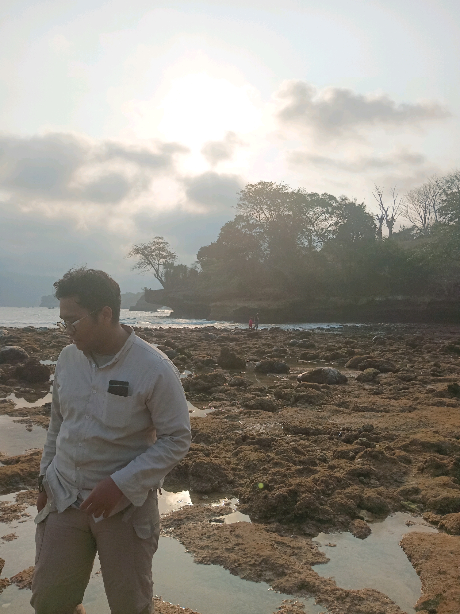 The image depicts a man standing on a rocky beach in Indonesia. The man is wearing a light-colored collared shirt and beige pants, with his hands in his pockets. He is looking down and to his left. The beach is covered in brown, textured rocks and small pools of water. In the background, the ocean is visible, with a gentle surf. Behind the ocean, there is a lush green hillside covered in trees. The sky is hazy and bright, suggesting either sunrise or sunset, with clouds obscuring the sun. Two small figures, possibly children, are visible walking near the water's edge in the mid-ground. The overall scene has a muted, natural color palette.