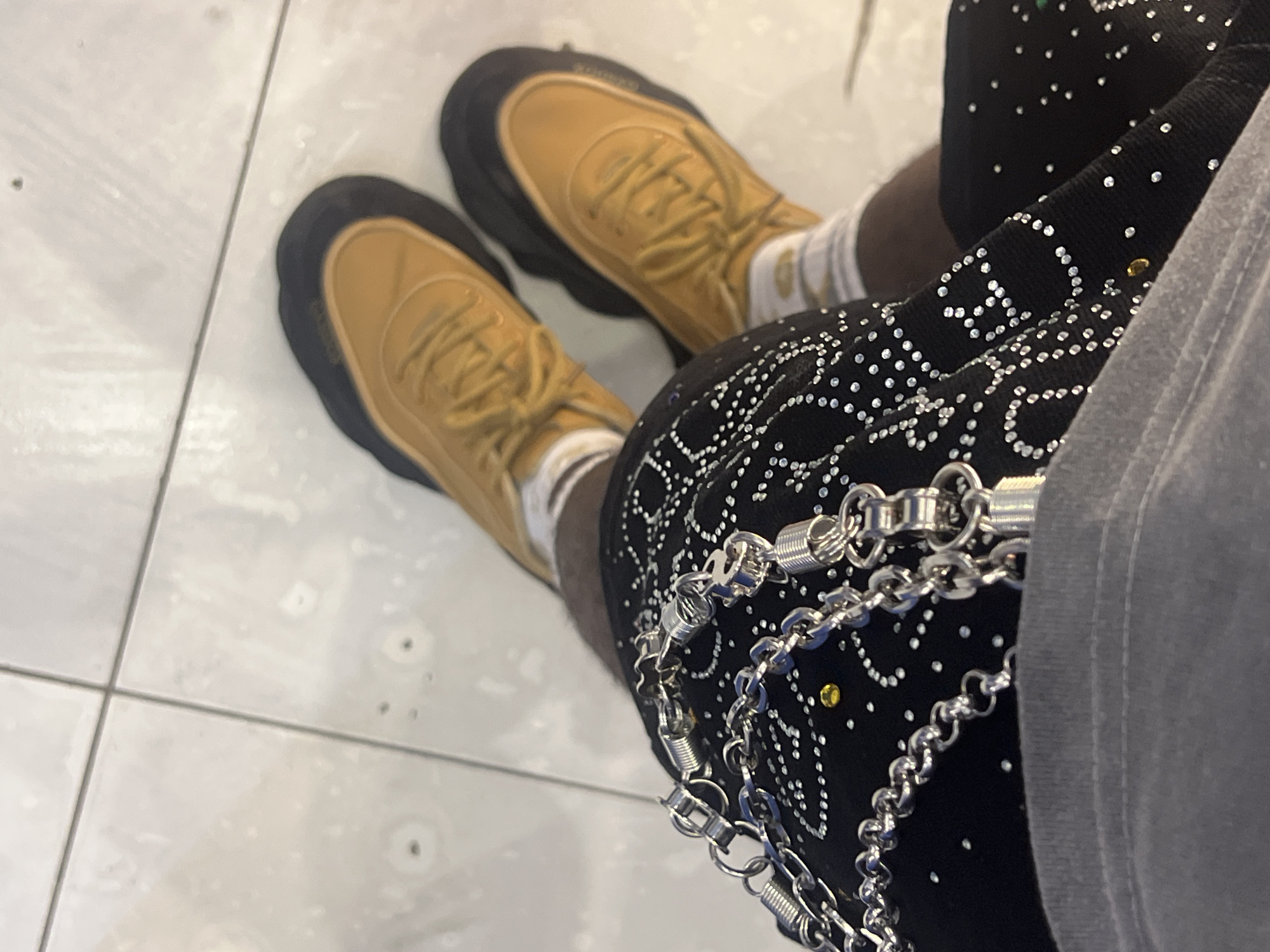 The image shows a person wearing mustard-colored sneakers with thick black soles, white socks, black shorts adorned with silver rhinestones, and silver chains. The shorts are also partially covered by a grey garment. The person is standing on a tiled floor. There is no other information that identifies the location as Howayi, Nigeria.