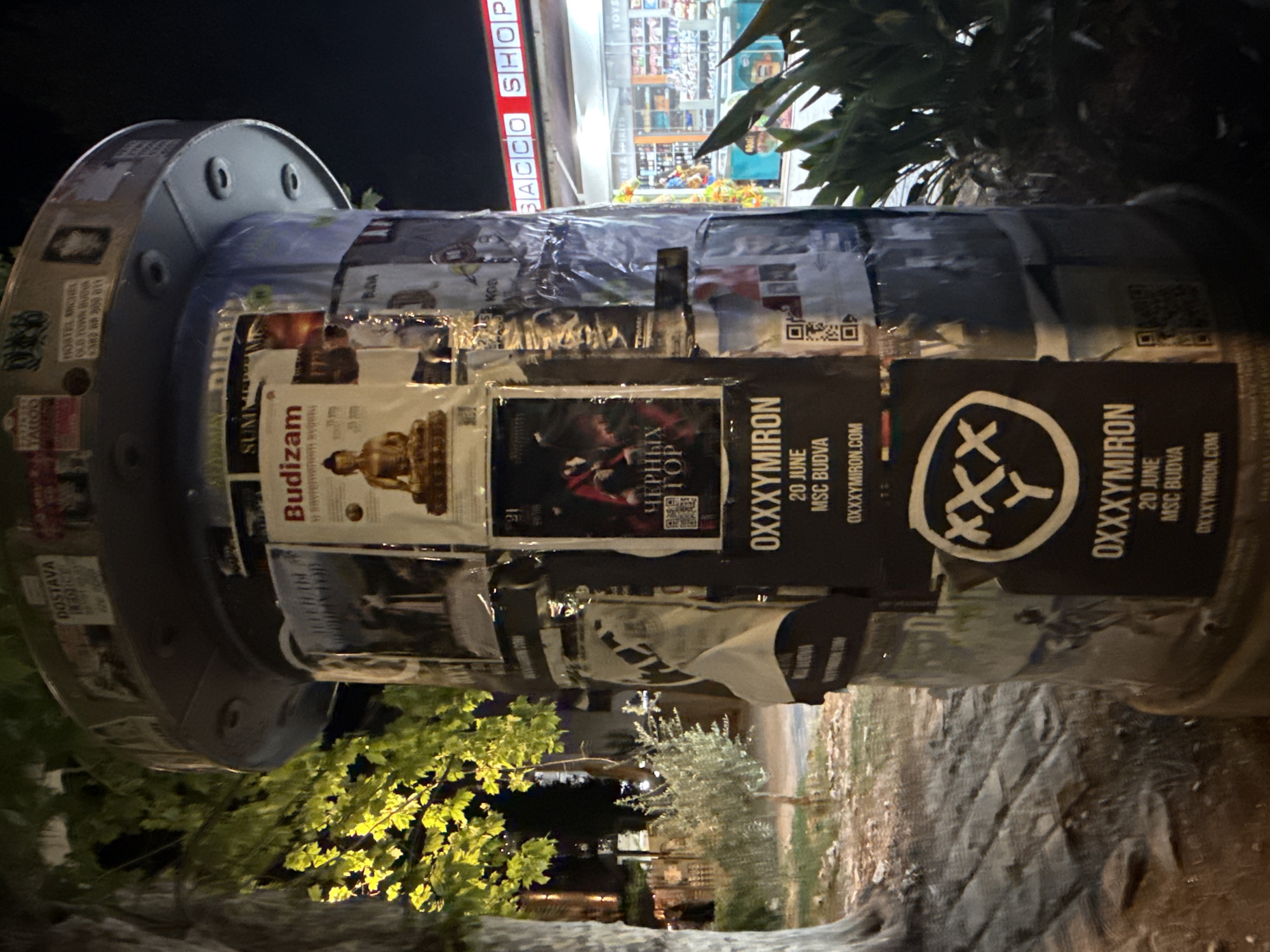 The image depicts a cylindrical public advertising pillar plastered with various posters and flyers in Budva, Montenegro. The pillar is situated outdoors at night, with a tree on the left casting shadows and illuminated foliage. In the background, a brightly lit shop with red signage that reads "BACCO SHOP" is visible, alongside rows of merchandise. The pillar itself is covered in numerous advertisements, some of which are torn or peeling. Prominent among them are posters related to "OXXXyMiroN" with dates and locations, and one featuring a Buddha image with the text "Budizam". QR codes are also present on some of the flyers. The ground appears to be paved with stone.