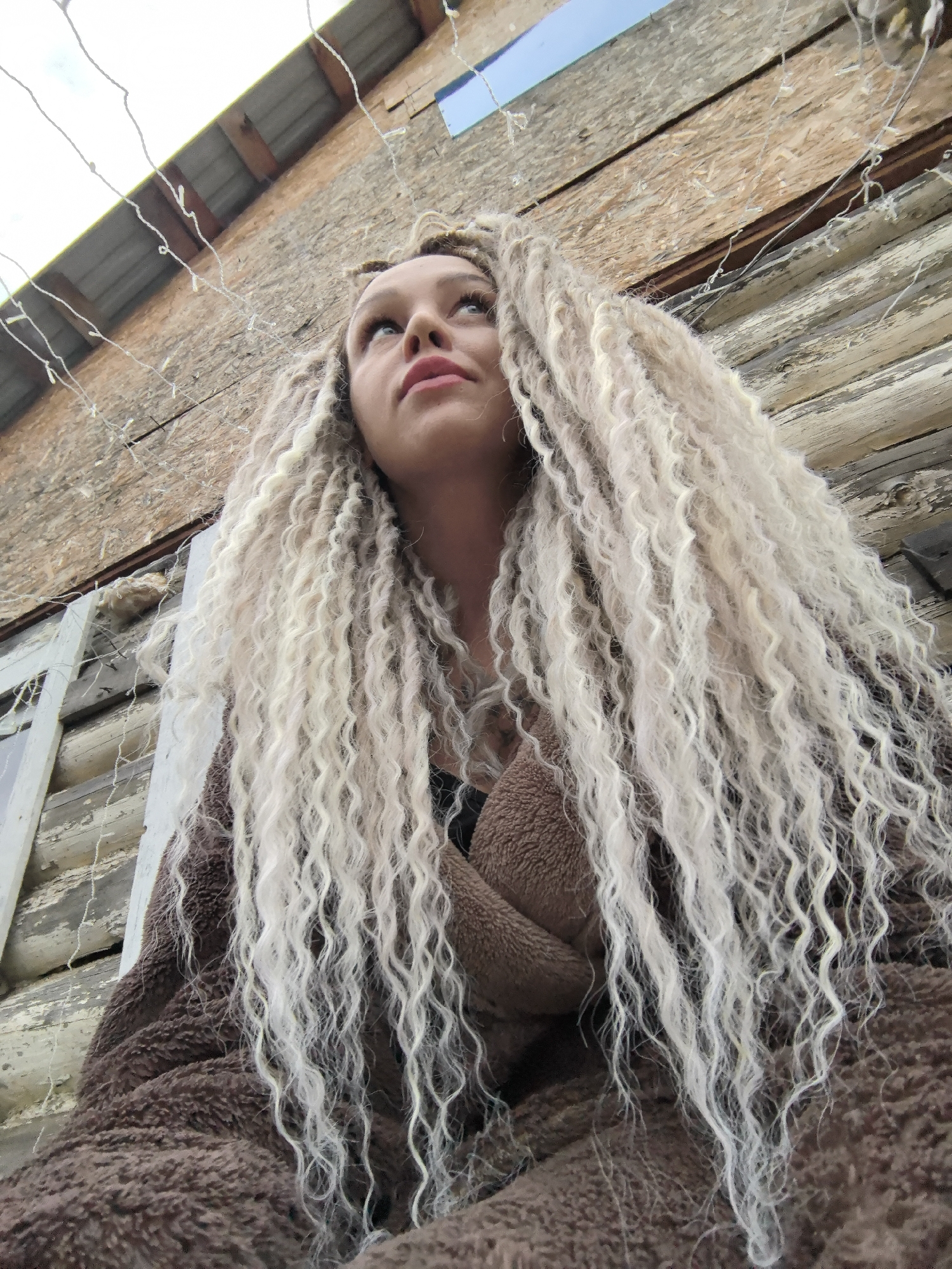 The image depicts a young woman with long, light-colored, curly hair. She is looking upwards and slightly to the left. She is wearing a brown, fuzzy garment. The background consists of a weathered, wooden structure and some string lights. A small patch of blue sky is visible at the top of the frame. The location and city of Noviy, Russia, cannot be confirmed based solely on the visual information provided in the image.