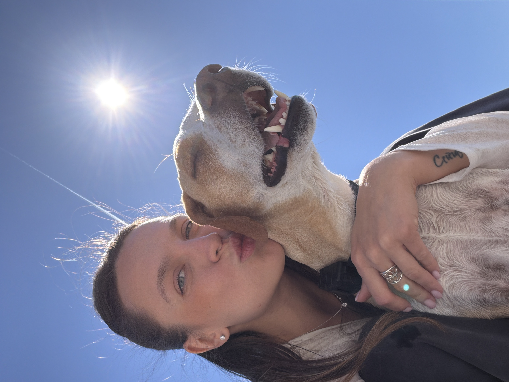 A young woman with long, dark hair and light eyes is holding a light-colored dog in her arms. She is making a kissing face, while the dog has its mouth open, seemingly smiling or yawning. The woman is wearing a silver ring on her hand, and a tattoo is visible on her arm. The background is a clear blue sky with the sun shining brightly. The location is outdoors, likely in Drobnići, Montenegro, given the request.