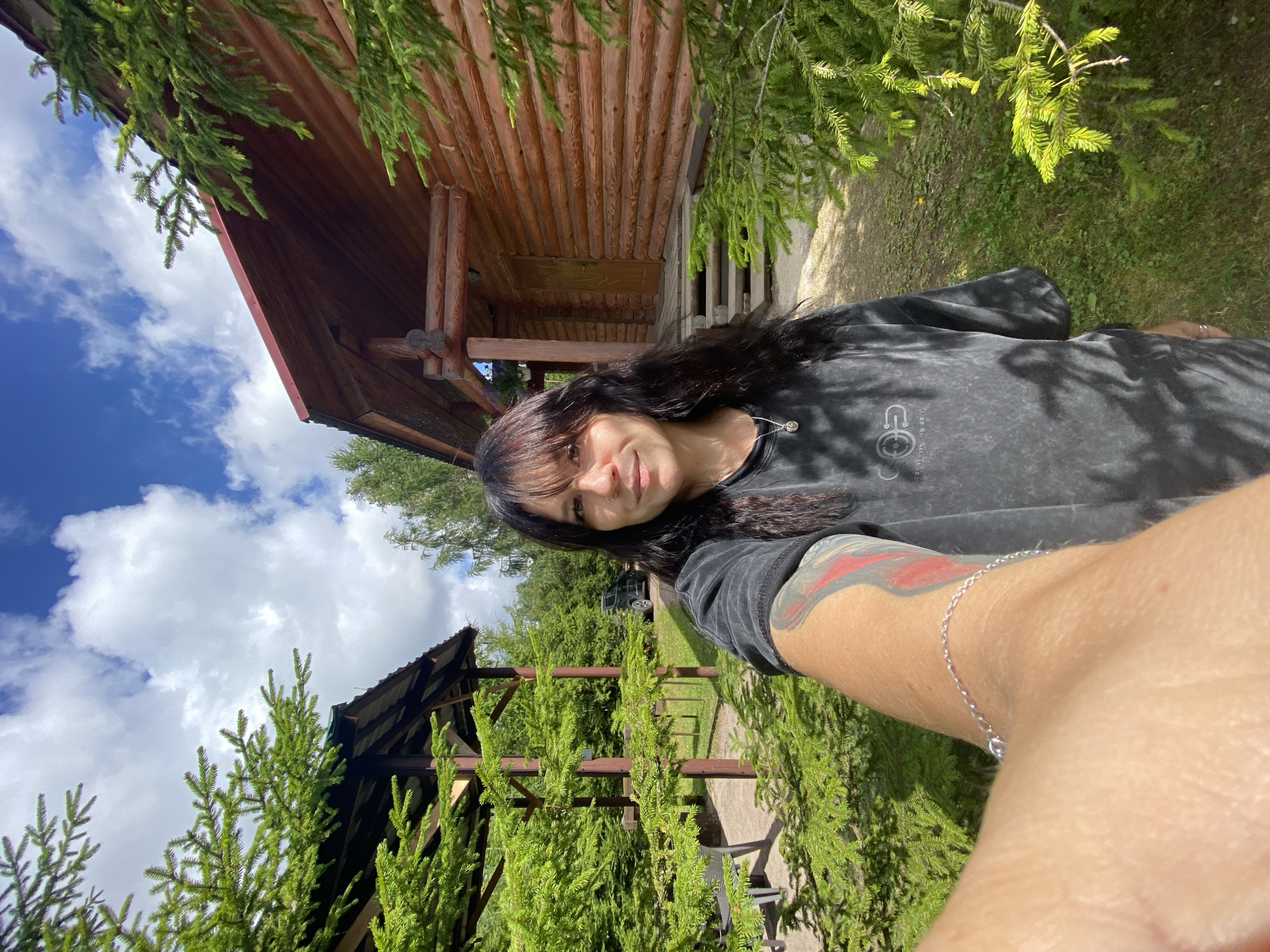 A woman with long dark hair and bangs, wearing a dark gray tie-dye t-shirt and a silver necklace, is taking a selfie outdoors. She has a tattoo on her left arm, which is visible along with a silver bracelet. She is standing in front of a wooden log house with a covered porch and steps leading up to the entrance. To the left of the house, there is an open-air gazebo with a table and chairs. The area is surrounded by lush green trees and grass, and the sky above is blue with white clouds. A black SUV is parked in the background to the left of the house. The scene appears to be in a rural or natural setting.