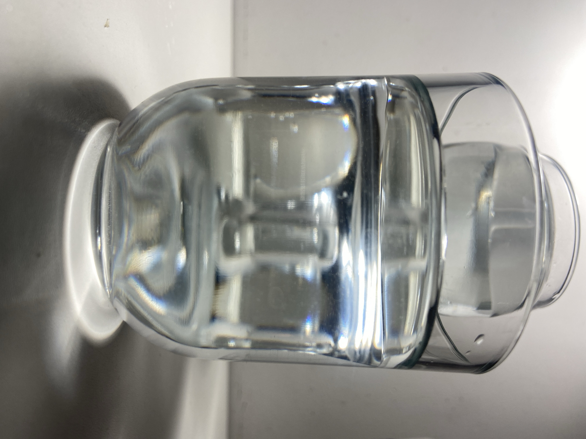 The image displays a close-up, low-angle view of a clear glass filled with water, resting on a flat, light-colored surface. Inside the larger glass, partially submerged in the water, is another smaller, clear glass or glass-like object, with its upper rim visible above the water line. The water appears clear, exhibiting optical effects such as light refraction and reflection, including subtle rainbow-like chromatic aberrations at various points. A strong, diffused light source from above illuminates the scene, creating bright highlights on the glass and water surface, and casting a distinct, dark, crescent-shaped shadow beneath the base of the main glass. The background is uniformly light-colored and out of focus. The image provides no identifiable geographical markers or contextual elements to determine its specific location within the city of Budva, Montenegro.