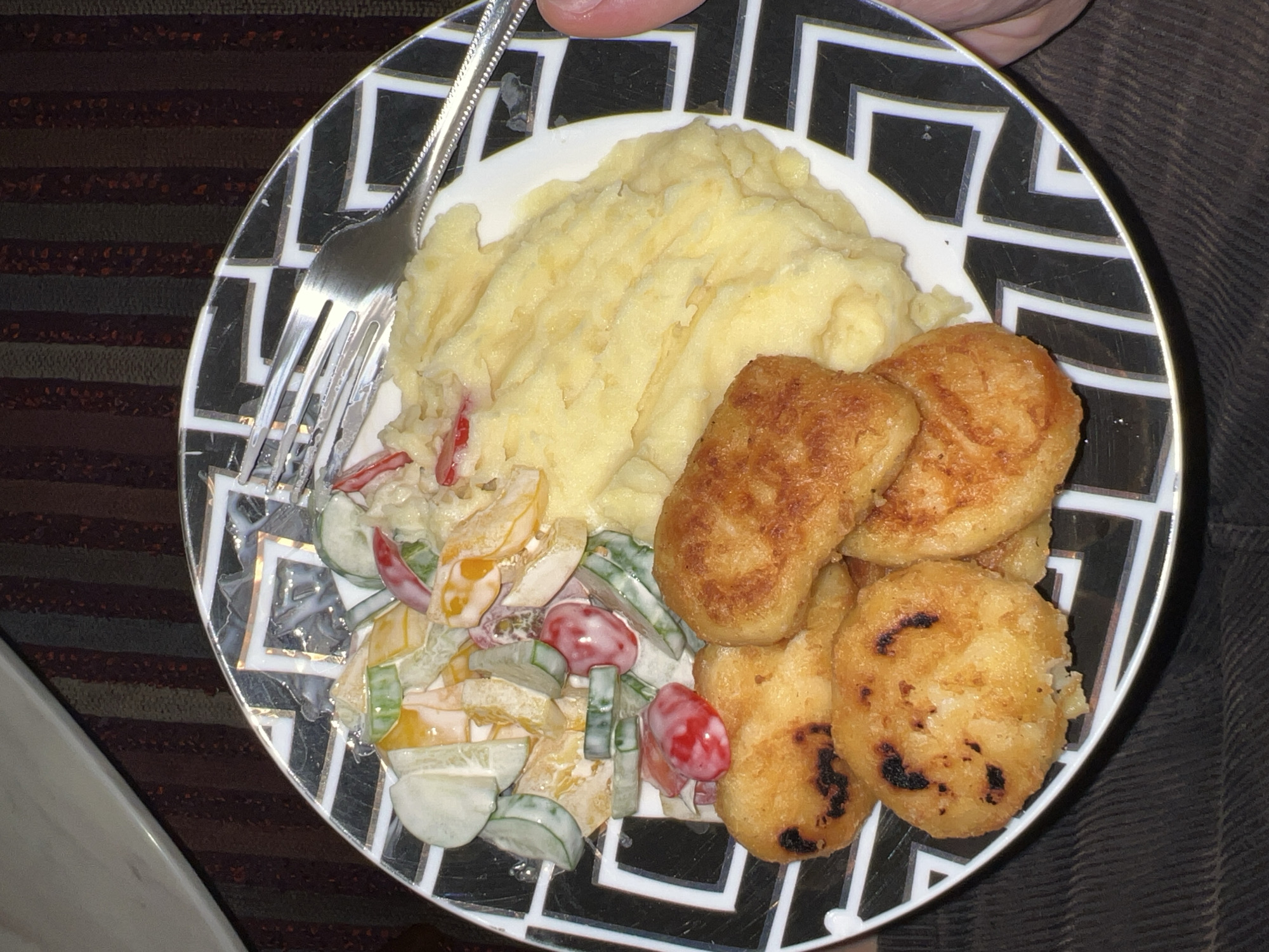A top-down, close-up shot depicts a plate of food on a surface with striped fabric and a person wearing brown clothing. The plate is black and white with a geometric, repeating square design. The food includes mashed potatoes, potato pancakes, and a creamy salad with tomatoes, cucumbers, and orange segments. A fork lies on the plate to the left of the mashed potatoes and salad. The location is Unknown, Unknown.