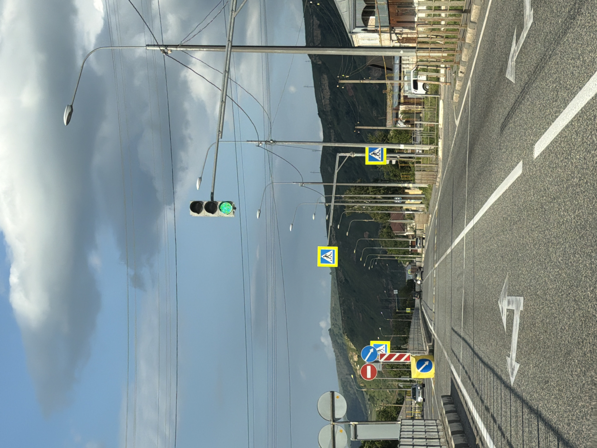 The image depicts a straight, paved road in Важное, Russia, with a green traffic light illuminated. On the left side of the road, there is a sidewalk bordered by a metal fence and some vegetation. Several traffic signs are visible, including a "no entry" sign, a blue circular sign with a white arrow indicating a turn, and a yellow square sign with a pedestrian crossing symbol. Further down the road, more pedestrian crossing signs and streetlights are present. The right side of the road features buildings and more metal fencing. The sky is partly cloudy with a mix of blue sky and grey clouds. The overall scene suggests a typical urban or suburban street environment.