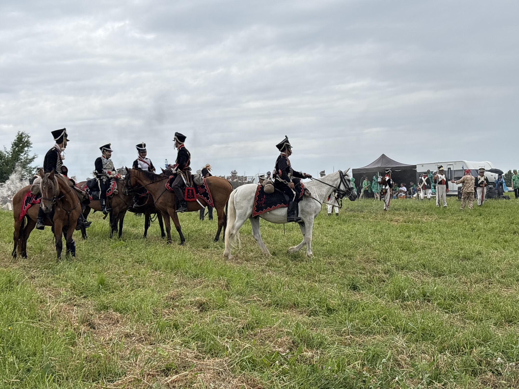 In a grassy field under a cloudy sky, a group of people in historical military attire on horseback are participating in a reenactment. Several men on horseback are prominently featured in the foreground, wearing elaborate uniforms with hats and decorative saddle blankets. One white horse stands out, its rider dressed in a dark uniform with red and gold trim. In the background, more figures on horseback and on foot are visible, along with tents, a trailer, and what appears to be smoke or steam rising from the left side of the frame. The event seems to be taking place in Goncharovo, Russia, given the location context.