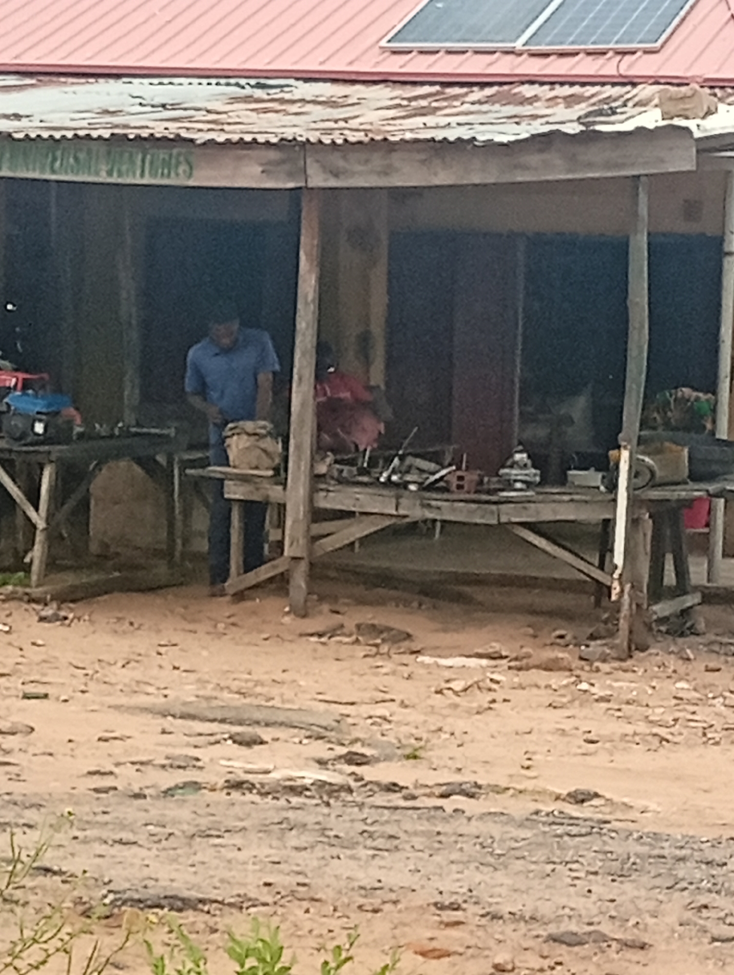 A medium shot captures an outdoor market stall in Gombe, Nigeria, characterized by a weathered corrugated metal roof and wooden supports. A man in a blue shirt and dark pants stands behind a table laden with tools and miscellaneous items, appearing to be working. On a table to his left, a blue and red power tool is visible. The ground is sandy and uneven, with debris scattered around. The backdrop suggests a simple, open-air establishment. The scene is illuminated by daylight, though a portion of the sky is obscured by the building.
