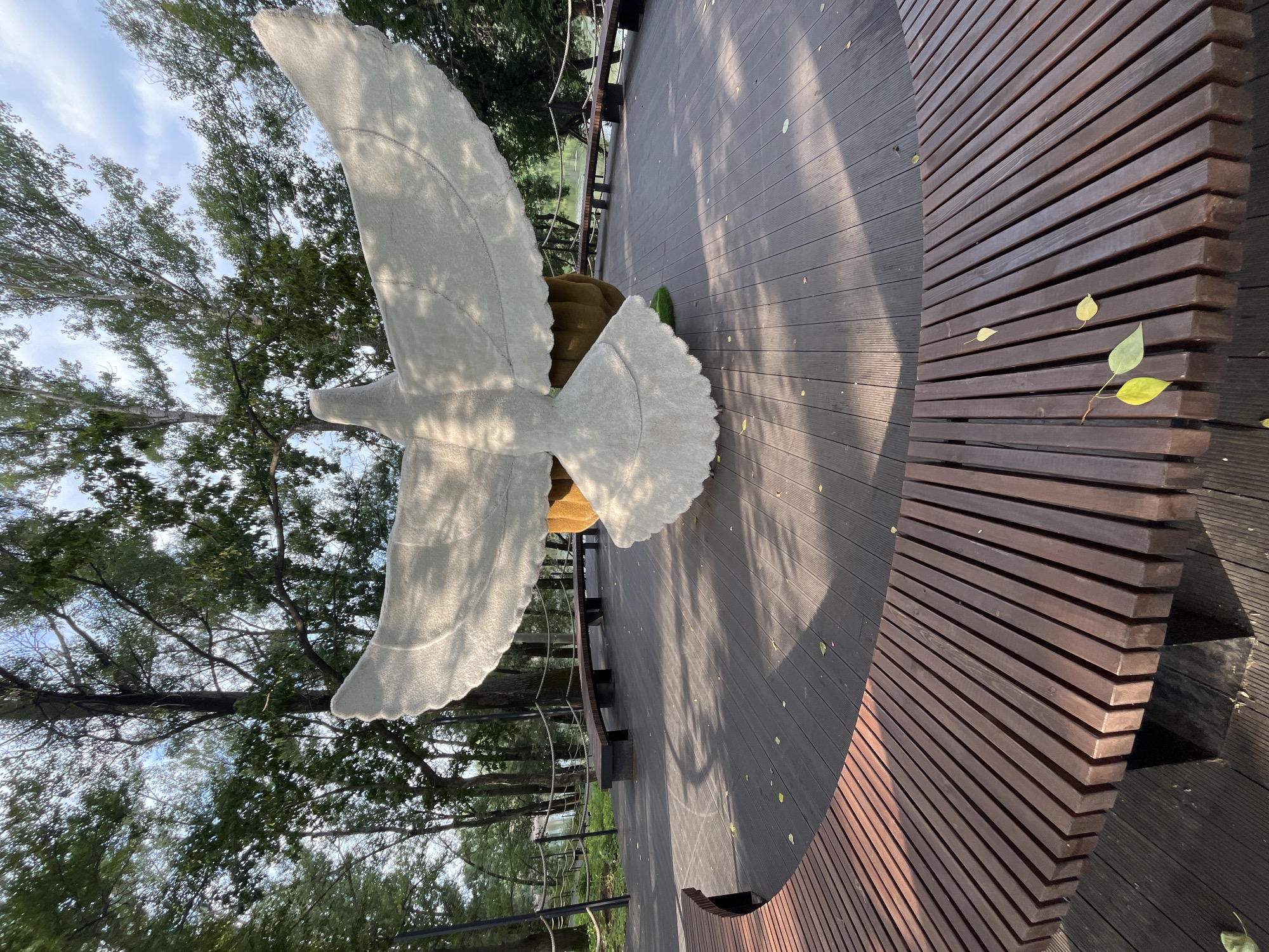 A white, stylized sculpture of a bird with outstretched wings is the central focus of the image, situated in an outdoor park or public space. The sculpture appears to be made of a textured material and rests on a large, bulbous, brown base. It is positioned on a circular wooden deck, which is surrounded by a curved wooden bench. The deck is made of dark wood planks, and scattered yellow and green leaves are visible on its surface and on the bench. In the background, a body of water, possibly a lake or river, is visible, bordered by a wooden railing and more trees. The scene is set amidst a backdrop of lush green trees under a partly cloudy sky. The overall atmosphere suggests a serene, natural environment within an urban setting. The sculpture is located in Blagoveshchensk, Russia.