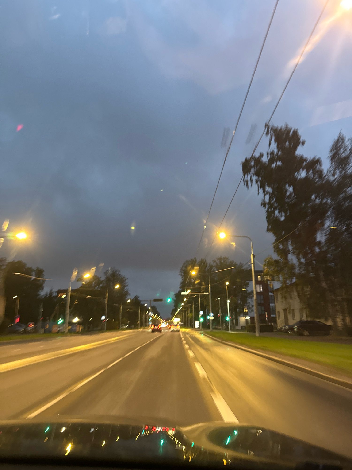 The image shows a view from inside a vehicle driving on a road in Tallinn, Estonia, at dusk or night. The road has multiple lanes and is illuminated by streetlights and car headlights. The sky is overcast and dark blue, with some clouds visible. Tall trees line the side of the road, and buildings can be seen in the background. There are other vehicles on the road, and traffic lights are visible. The reflection of streetlights and the car's dashboard can be seen on the windshield. The overall impression is that of a typical urban driving scene in cool weather.