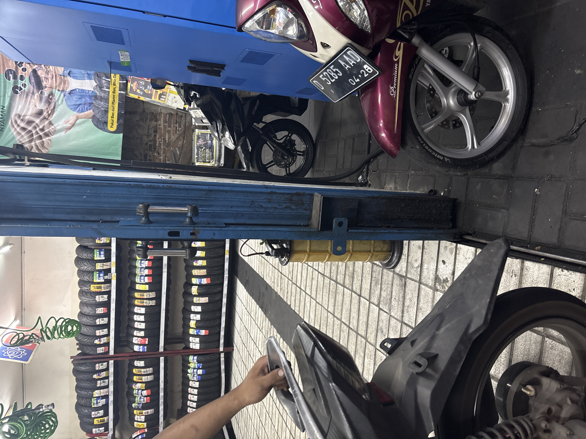 In North Jakarta, Indonesia, a motorcycle tire shop is depicted. The scene features multiple shelves stocked with various motorcycle tires, some with price tags attached. In the foreground, a person's hand is visible, holding onto the back of a motorcycle. Two motorcycles are parked side-by-side, with one in the foreground partially obscuring the other. To the right, a large blue cabinet or machine stands, and a yellow oil drain can is positioned between the two motorcycles and the blue cabinet. A banner with text and an image of a man and tires hangs on the wall behind the motorcycles. The floor is tiled with a light gray and white pattern, and some dark streaks are visible on the ground. The overall impression is of a busy service or retail environment for motorcycles and their parts.