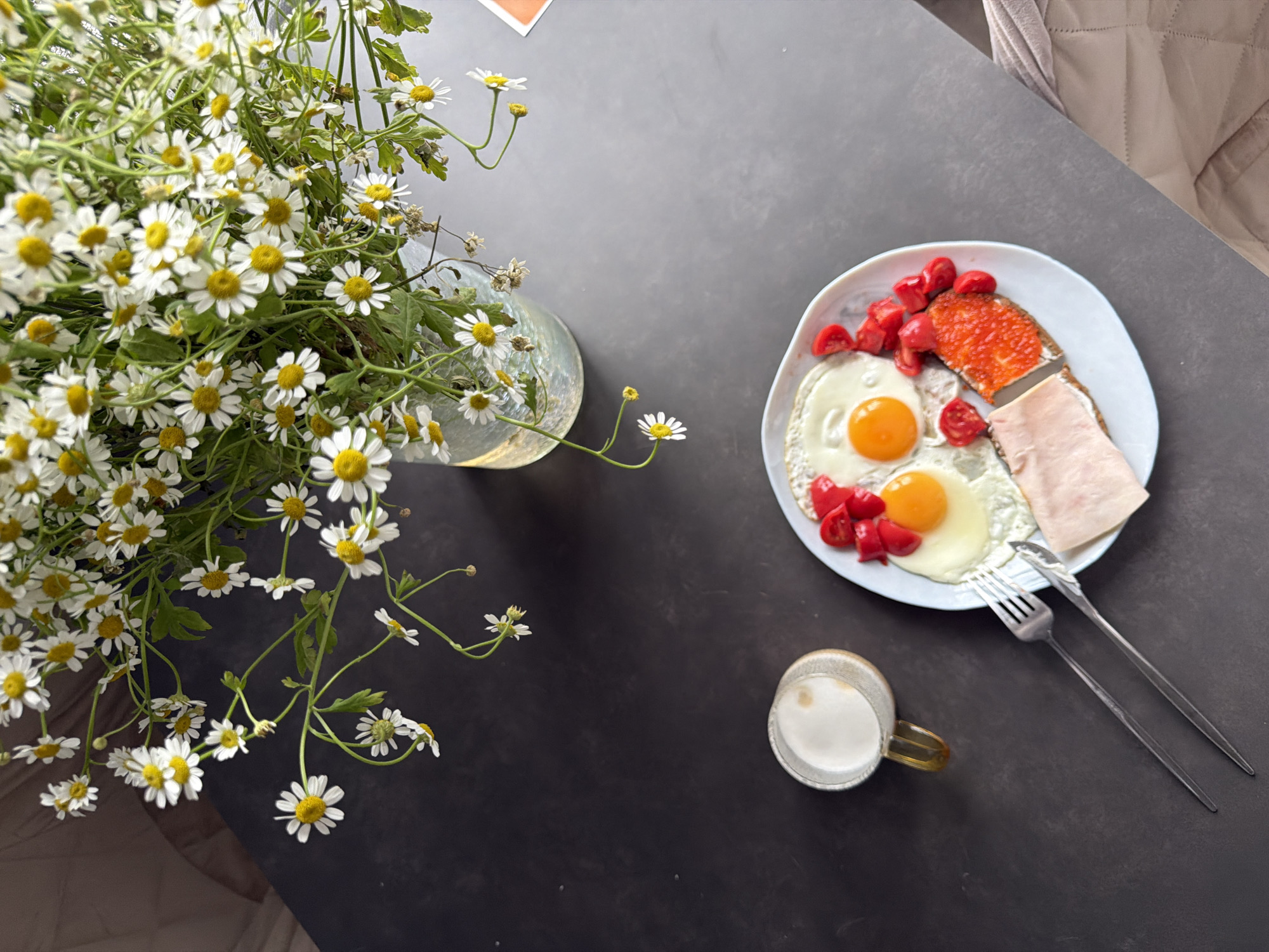The image depicts a still life arrangement on a dark gray or black surface. To the left, a large bouquet of white daisies with yellow centers is arranged in a translucent vase with an opalescent sheen. The daisies are in full bloom, with some stems extending outwards from the vase.
To the right, a light blue plate holds a breakfast meal. The meal consists of two fried eggs with bright yellow yolks, chopped red tomatoes, two slices of bread, one topped with orange caviar and the other with sliced ham. Beside the plate, a silver fork and knife are laid out.
Below the plate is a small, clear glass mug containing a white liquid, presumably milk or a milky beverage. The mug has a yellow tinted handle. The arrangement is set against a textured backdrop in the upper right corner, which may be a piece of furniture or fabric.
There are no visible people or indications of a specific city or location.