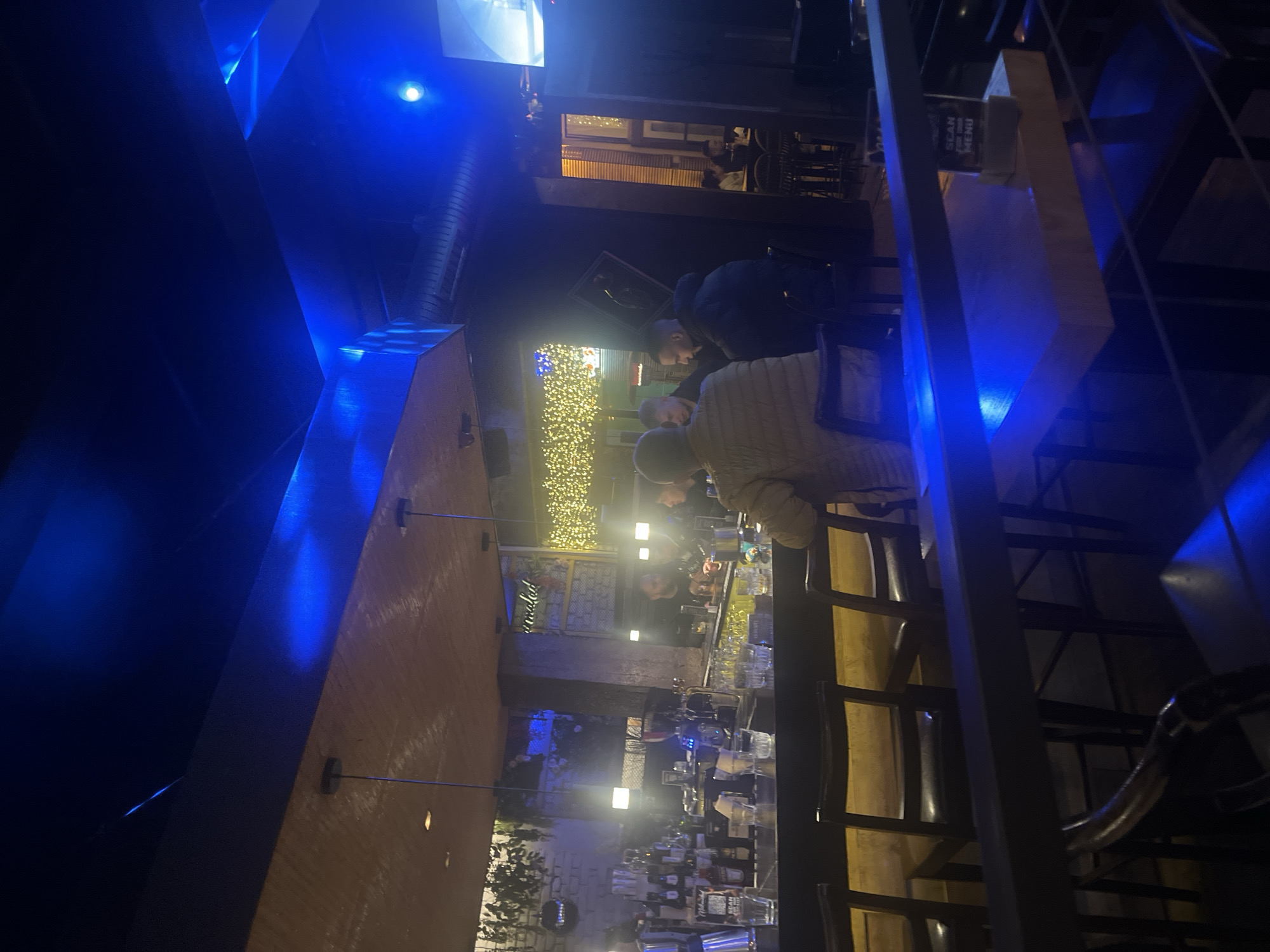 The image shows the interior of a bar or restaurant. Several people are seated at the bar, which is illuminated with warm lighting. Bottles and glasses line the bar's surface. A person is sitting and leaning over the bar in a tan shirt. The background features a decorative wall with string lights, and a plant. The bar is in Plovdiv, Bulgaria. The bar also has a large blue lit structure on the ceiling.