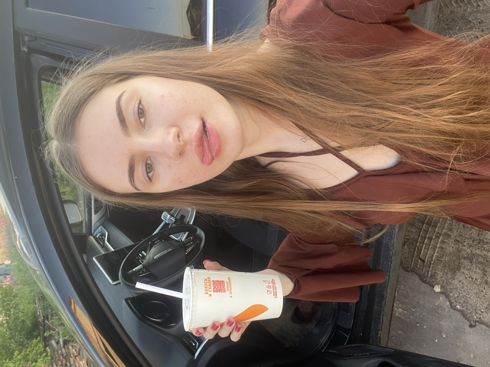 A young woman with long, blonde hair is standing next to the open driver's side door of a black car. She is holding a white paper cup with a straw in her right hand, and her tongue is sticking out of her mouth. The cup has text and a QR code on it. The car's interior, including the steering wheel and dashboard, is visible. The background shows greenery and some stacked materials. The ground appears to be a paved surface. The woman is wearing a rust-colored top with a low neckline. The image seems to be taken outdoors during daylight. The provided metadata suggests the location is Медведево, Russia.