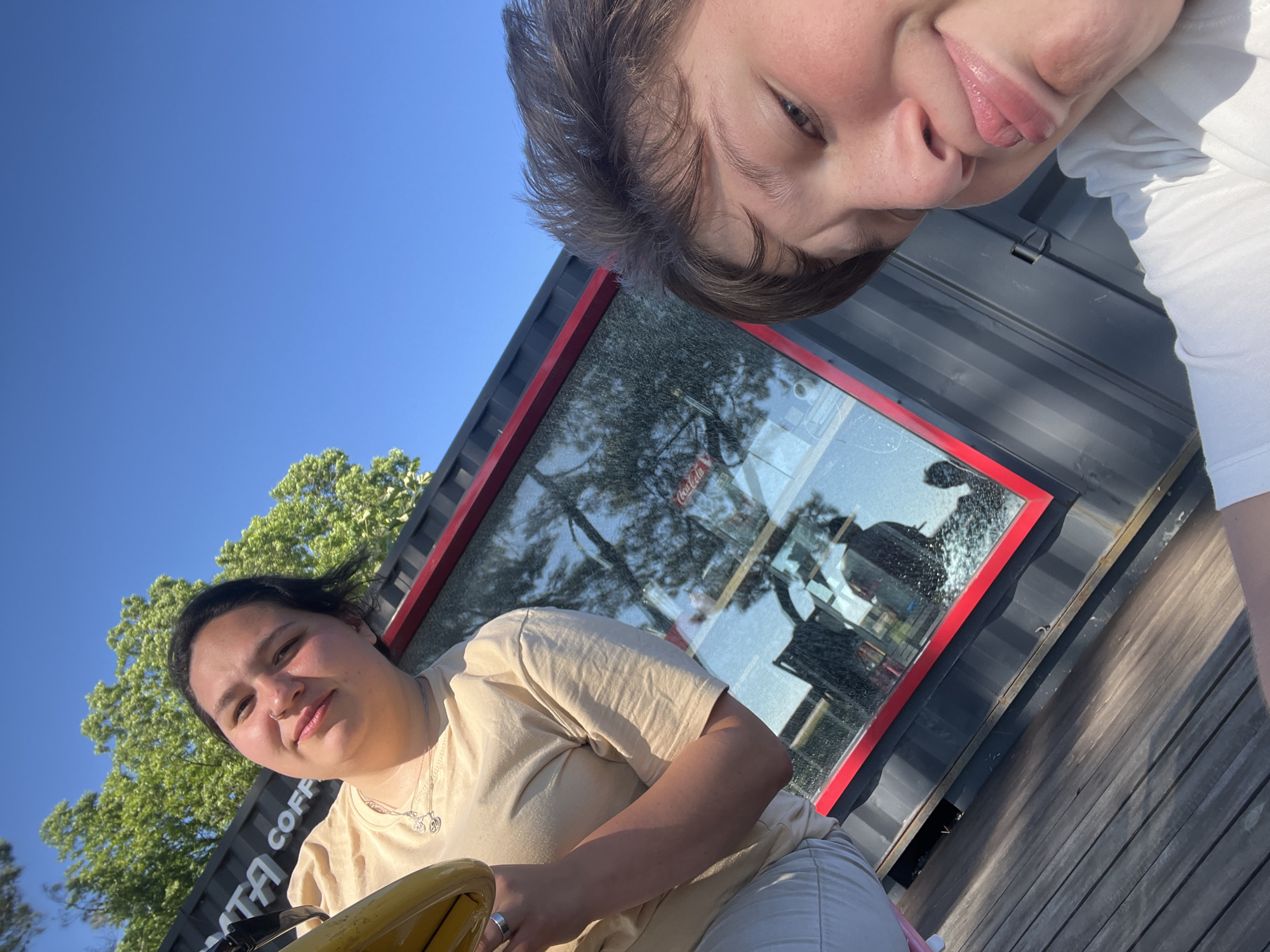 This media file features two young individuals, likely friends, posing for a selfie outdoors. The first person, in the foreground and to the left, is a young woman with dark hair pulled back and a light tan t-shirt. She is looking directly at the camera with a slight smile. She is wearing a necklace with a charm and a ring on her left hand. The second person, partially visible in the upper right of the frame, has brown hair and is looking down at the camera. They are wearing a white t-shirt.
The scene appears to be a sunny day, with a clear blue sky visible. They are standing near a dark, modular structure with a red trim around a large window. Through the window, reflections of trees and sky are visible, along with what appears to be a Coca-Cola logo and some interior elements, suggesting it might be a kiosk or a small building. The ground beneath them is made of wooden planks. The overall mood seems relaxed and happy, suggesting a casual outing.
Visible text includes "COFF" on the side of the dark structure, implying it may be a coffee stand or shop. The image appears to be taken in Ochamchire, Abkhazia. The sun is casting long shadows, indicating it might be late afternoon.