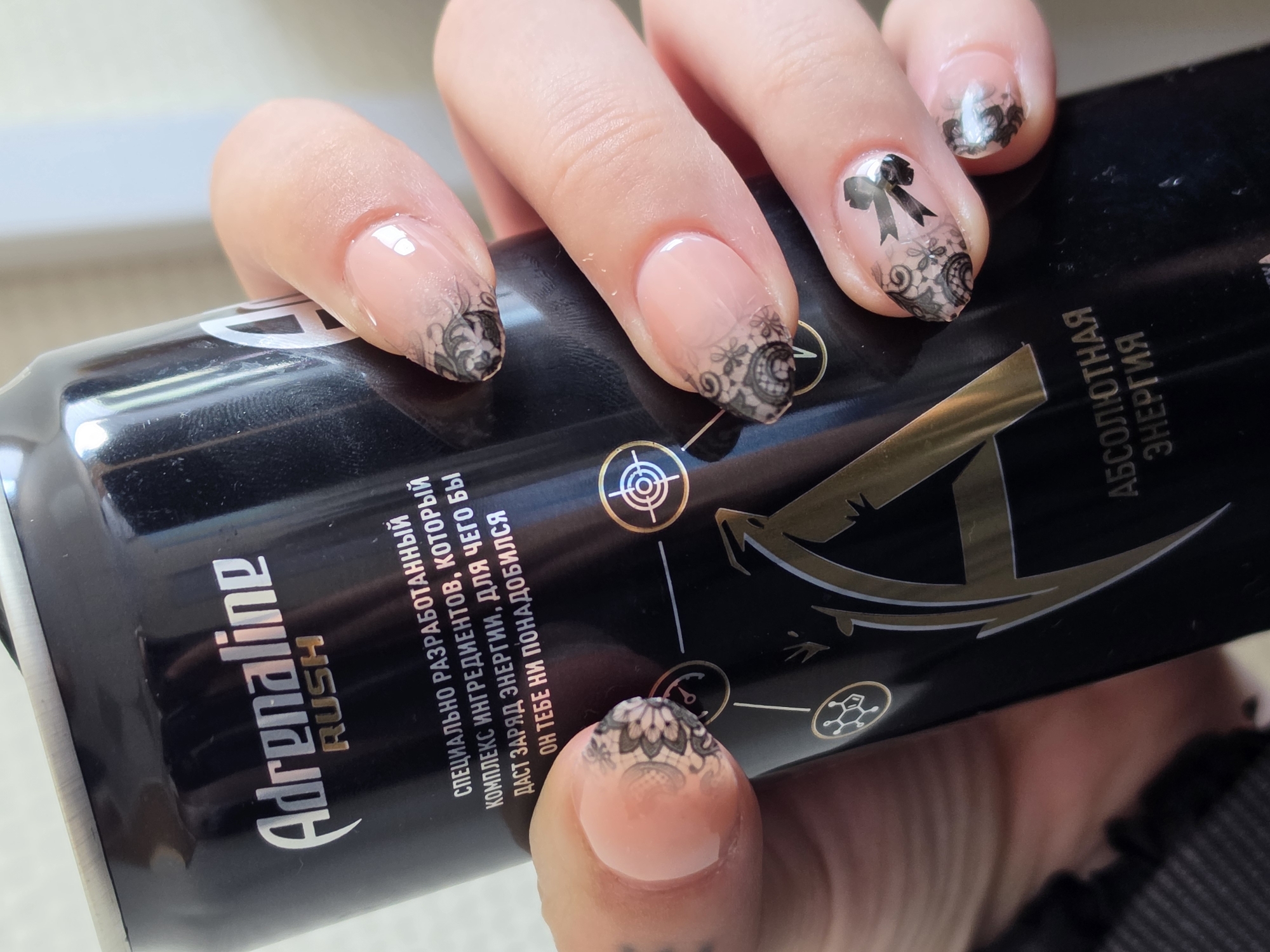 A close-up shot shows a person's hand holding a black can of Adrenaline Rush energy drink. The can features the brand's logo and text in Russian. The person's nails are painted with a nude base coat and adorned with intricate black lace patterns and a small black bow on one nail. The background is blurred, but appears to be indoors. There is no specific information in the image to determine the location in Nizhny Novgorod, Russia.