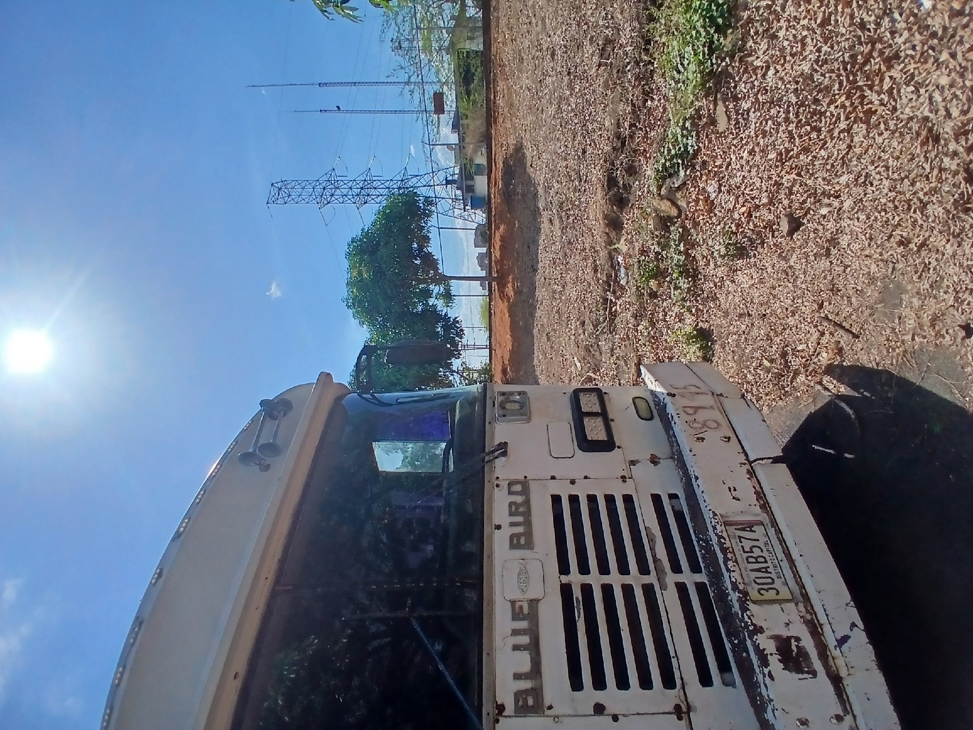 The image captures a bright, sunny outdoor scene in Ciudad Guayana, Venezuela, featuring a large, weathered white industrial vehicle in the foreground. The vehicle, possibly a heavy-duty truck or specialized utility vehicle, is viewed from its front-left side. Its prominent features include a vertical front grille, a visible square headlight on the right, and two air horns mounted on the roof. The brand name "BLUE BIRD" is embossed on the front panel, alongside a logo. A license plate displaying "30AB57A" is attached to the front, with the partially visible text "DISTRIT CAPITAL" beneath it.
In the mid-ground and background, a clear blue sky dominates, with a bright sunflare visible on the left side. Several tall metal structures, appearing to be electricity transmission or communication towers, rise into the sky, including a notable lattice tower. Green trees are interspersed among these structures. The ground to the right of the vehicle is unpaved and appears dry, covered with reddish-brown soil, small stones, and dry leaves, with a strip of green vegetation bordering it. No people are visible in the scene. The overall impression is that of a utility or industrial area on a clear, dry day. The vehicle appears stationary, and the bright light suggests it is daytime.