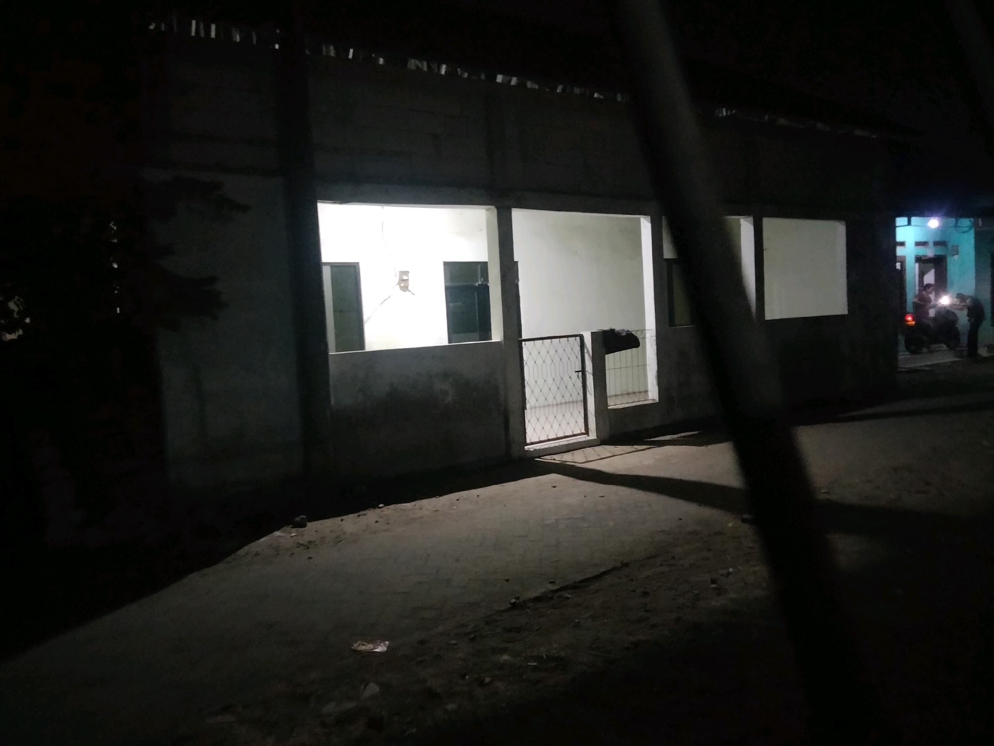 The image shows an exterior view of a building at night in Suka Sari, Indonesia. The building appears to be a single-story structure with a rough, unfinished concrete facade and a corrugated roof. Several large, open doorways or windows reveal illuminated interiors. In the foreground, the ground is dark asphalt or concrete, with some scattered debris.
On the right side of the image, further down the street, another building with a bright blue facade is visible, with its entrance lit. Two individuals are standing near a motorcycle in front of this blue building, and a third person is partially visible. The lighting suggests it is late evening or nighttime. The overall impression is a somewhat unkempt, dimly lit urban or semi-urban environment.