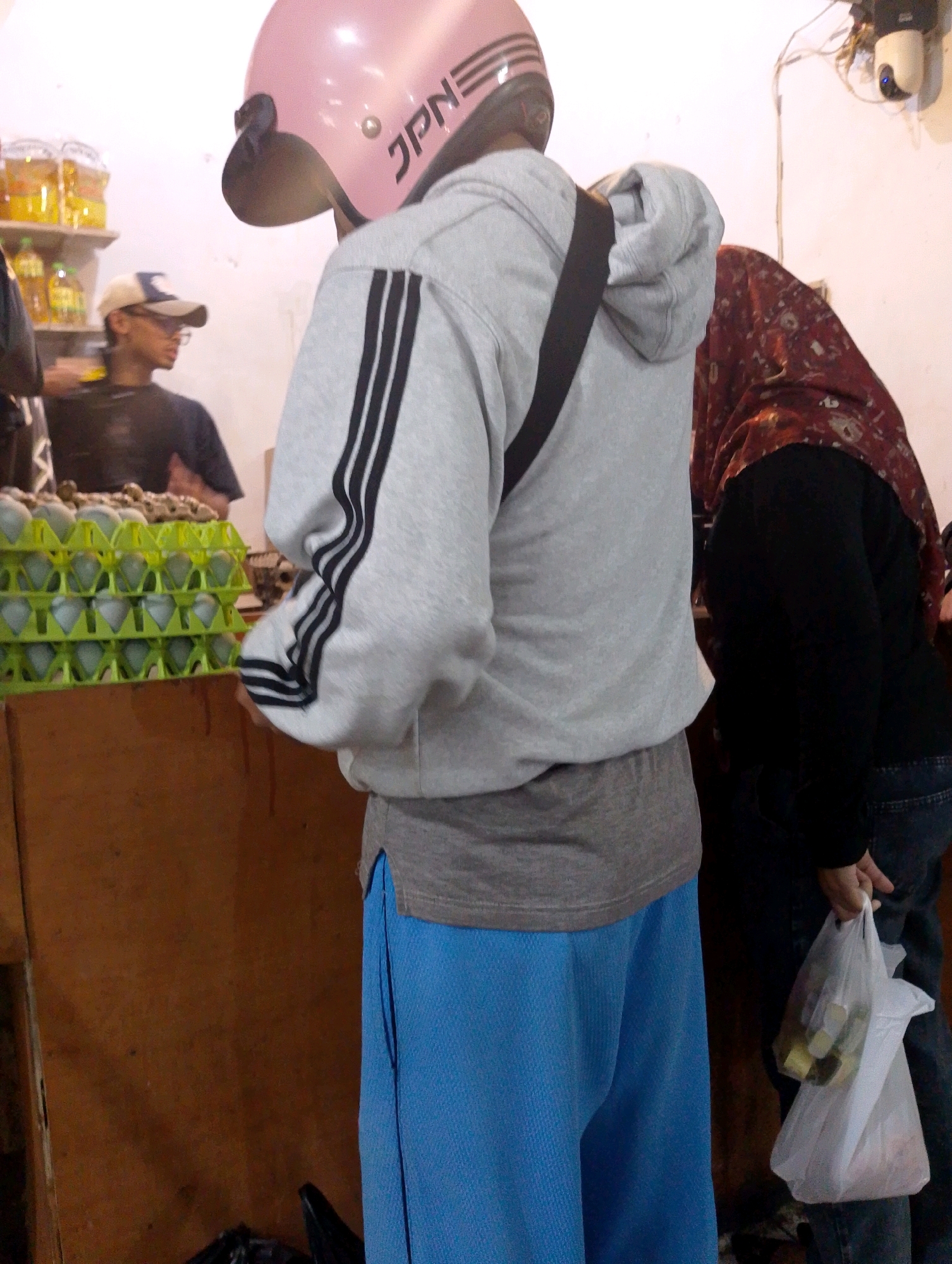 The image shows a market stall in Lembang, Indonesia. In the foreground, a person wearing a pink helmet, grey hoodie with black stripes, and blue pants is standing. They are partially obscuring a display of green egg cartons filled with eggs. Behind this person, another individual wearing a red and white patterned headscarf and a black top is visible, holding a white plastic bag containing items. In the background, shelves are stocked with bottles, and a surveillance camera is mounted on the wall. The scene suggests a busy marketplace environment.